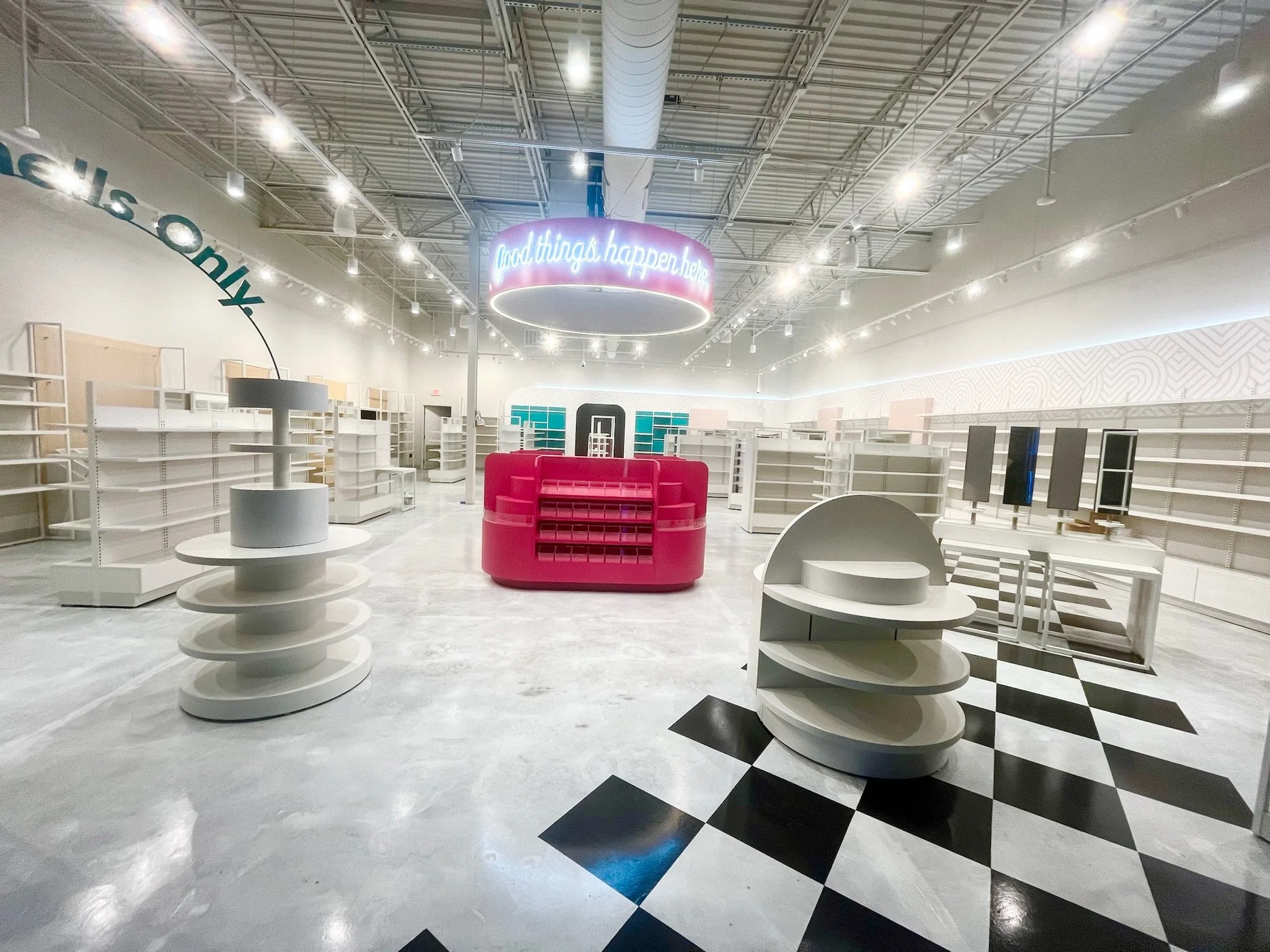 Empty retail store with white shelves, a pink display, and a neon sign that says "good things happen here" hanging from the ceiling. The store appears to be closed or under renovation.