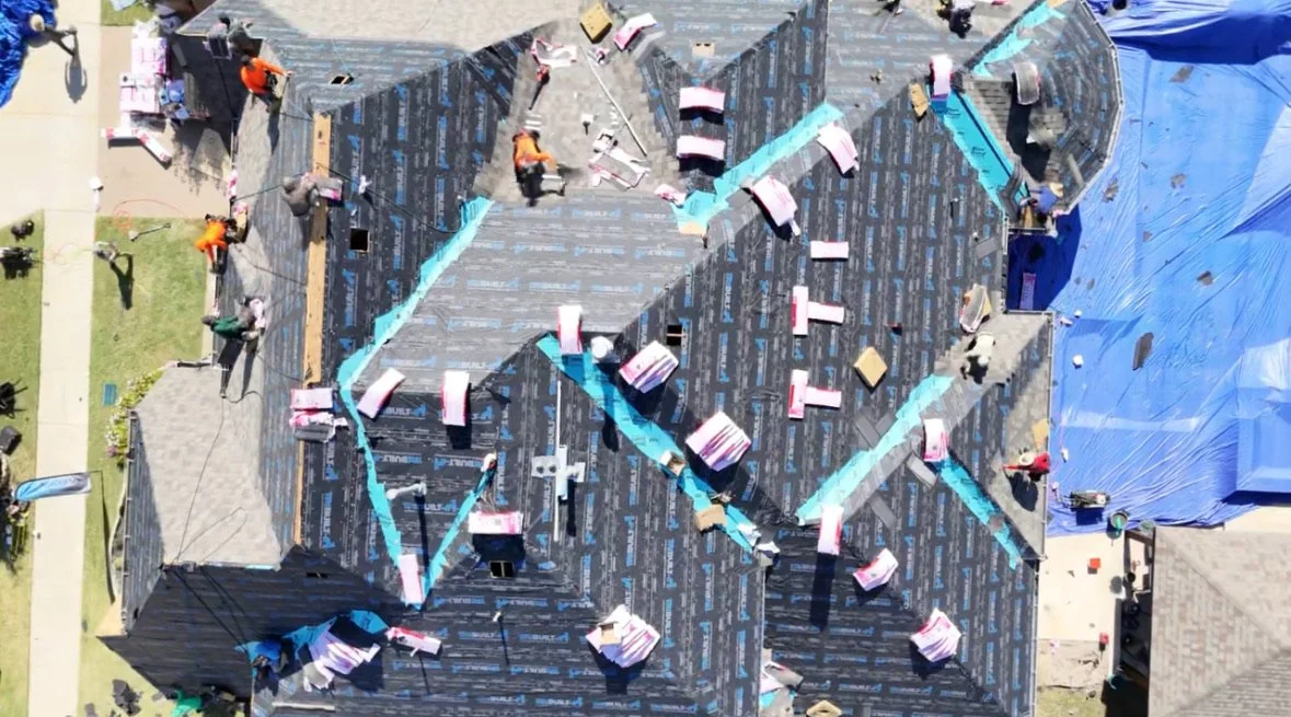 Aerial view of a house under construction with workers installing roofing; the roof is partially covered with foam and roofing materials.