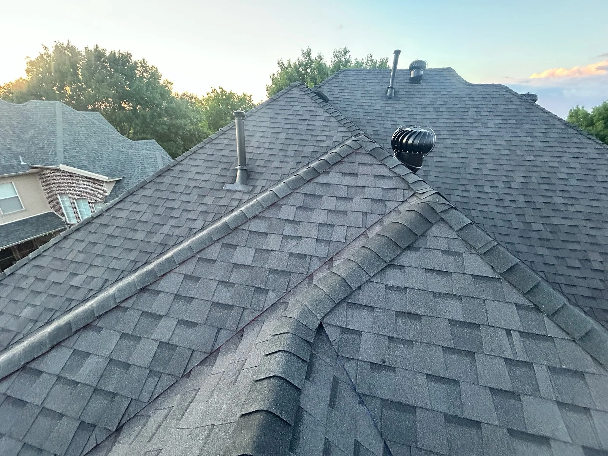 View of multiple rooftops with shingled roofs, some with vents, under a partly cloudy sky with trees in the background.
