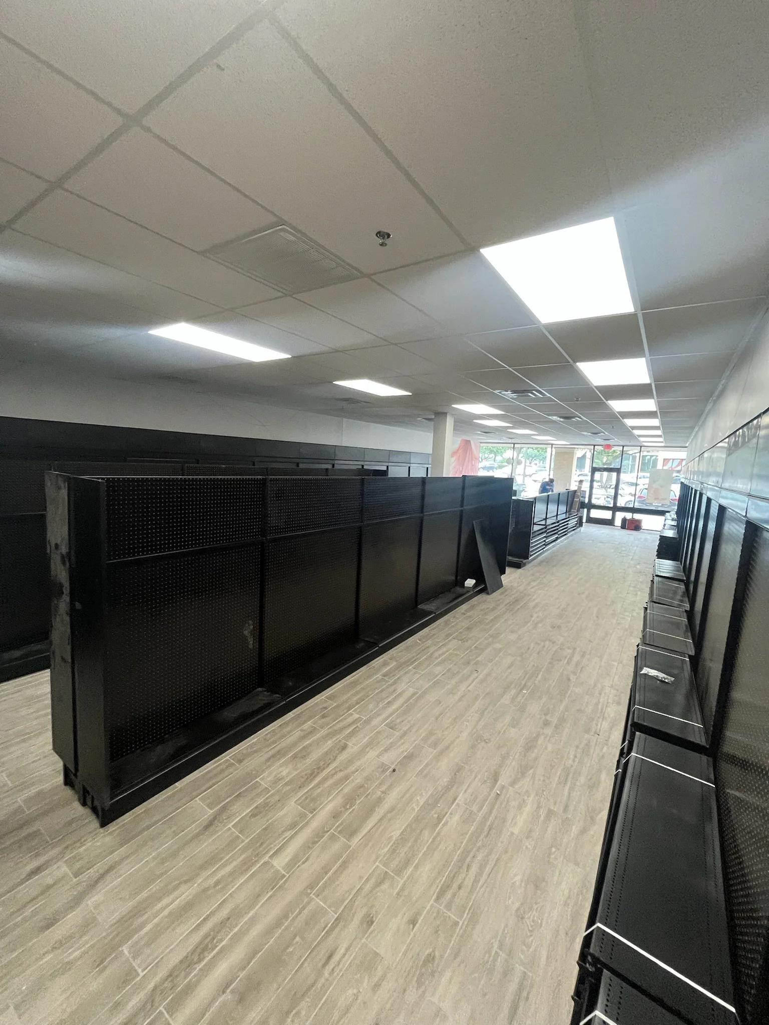 Empty retail space with black shelving units, a row of black chairs, white ceiling tiles, and a large glass entrance with visible parking lot outside.