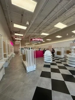 Empty retail store with black and white checkered flooring, white display shelves, and a pink counter in the background. Overhead lighting and a ceiling with exposed pipes and vents.