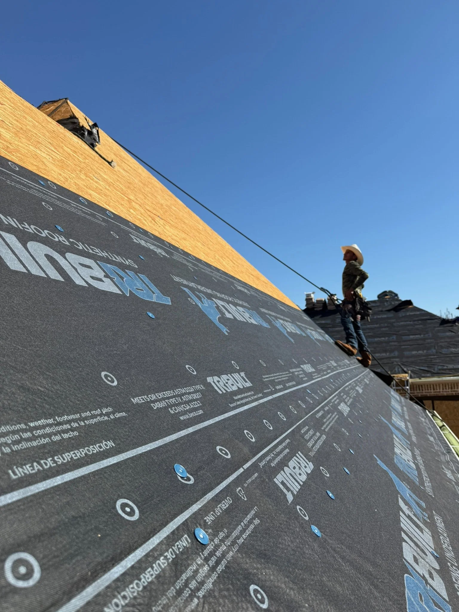 A construction worker standing on a roof under construction, with a clear blue sky in the background. The roof has black underlayment and wooden sheathing, and the worker is wearing a cowboy hat.