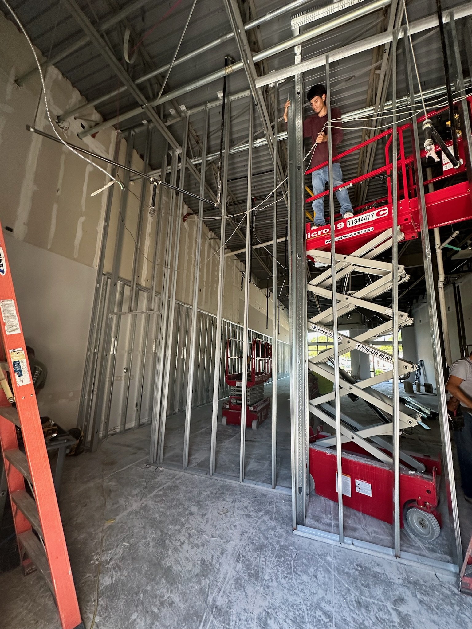 Inside a building under construction with metal framing, electrical wiring, and a worker on a man lift. The room has unfinished walls and concrete floors.