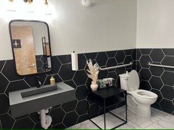 Bathroom with black hexagon tile walls, white ceiling, and light-colored floor. Features a gray floating sink with a mirror above, a small table with decorative items, and a white toilet.