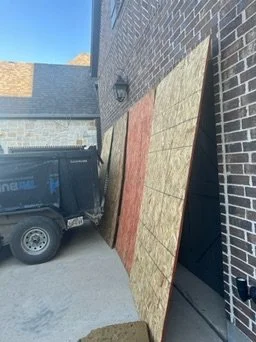 Two large sheets of plywood leaning against a brick wall, with a black utility trailer nearby under a clear blue sky.