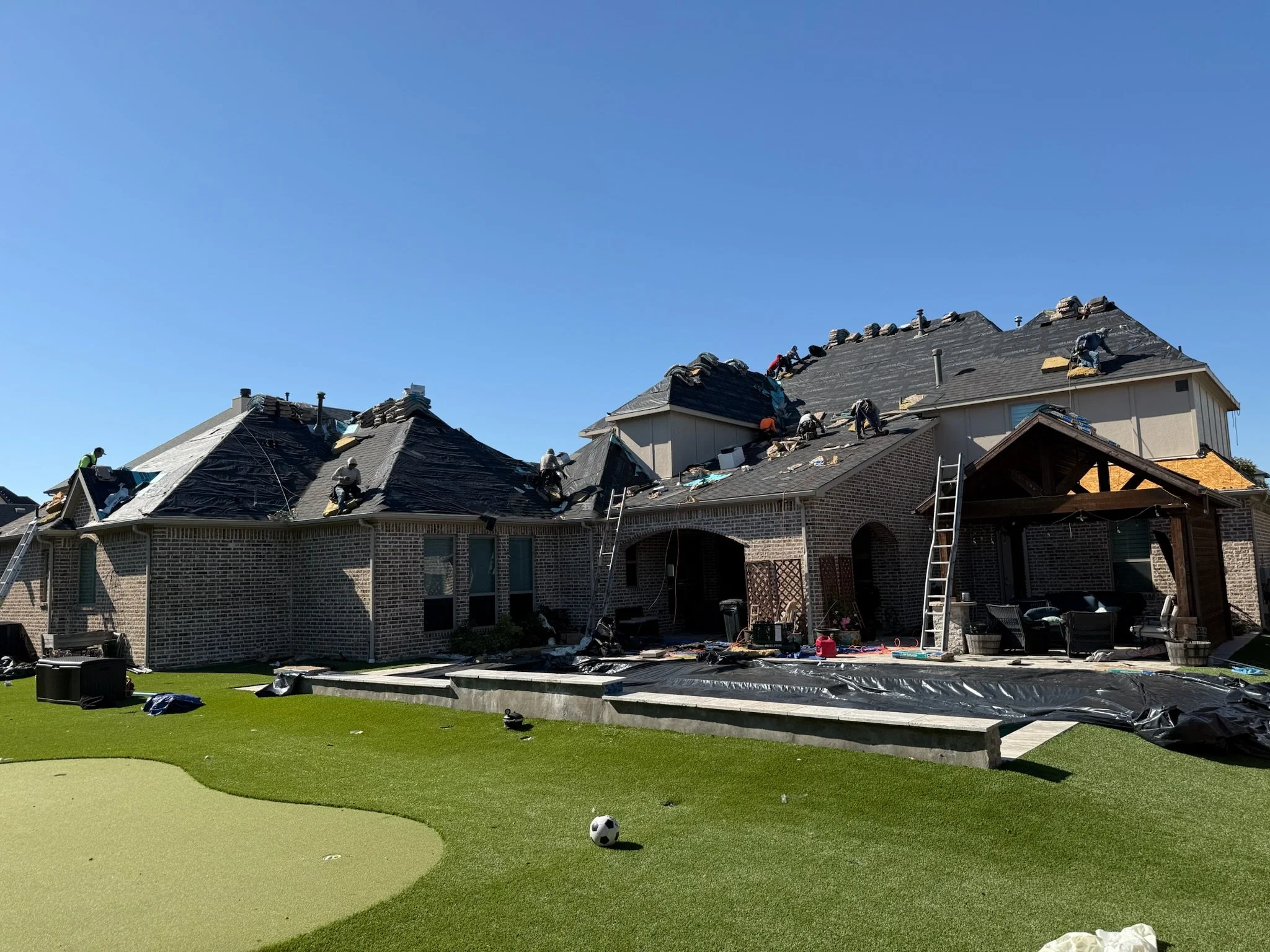 House with workers on its roof renovation under a clear blue sky, with a green backyard featuring a small soccer ball.