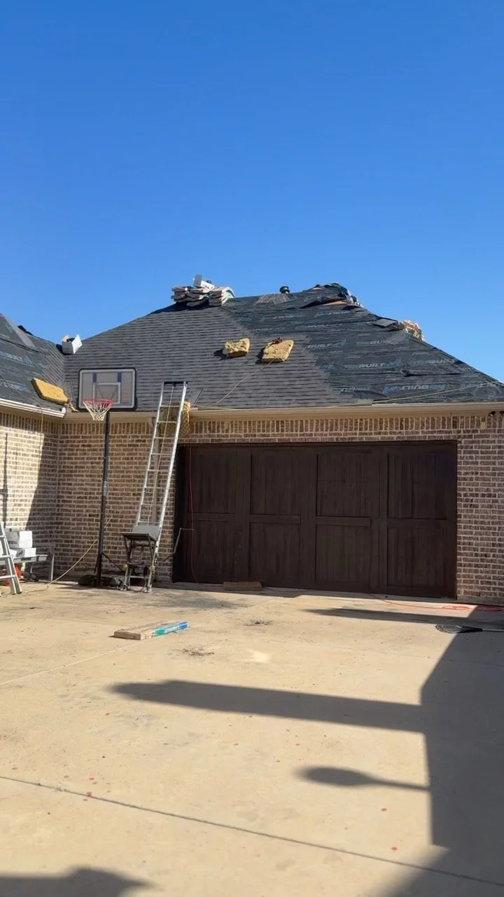 House with a brick exterior undergoing roof repairs, visible under construction tools and materials, with a basketball hoop and ladder nearby, outside on a clear day.