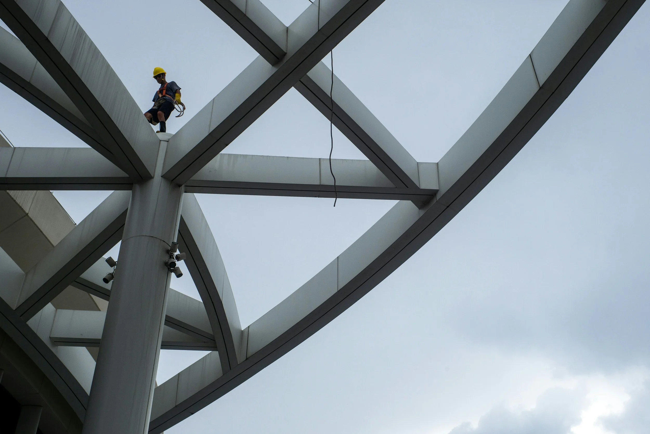 A construction worker wearing a yellow safety helmet and harness working on a modern white architectural structure with steel beams against a cloudy sky.