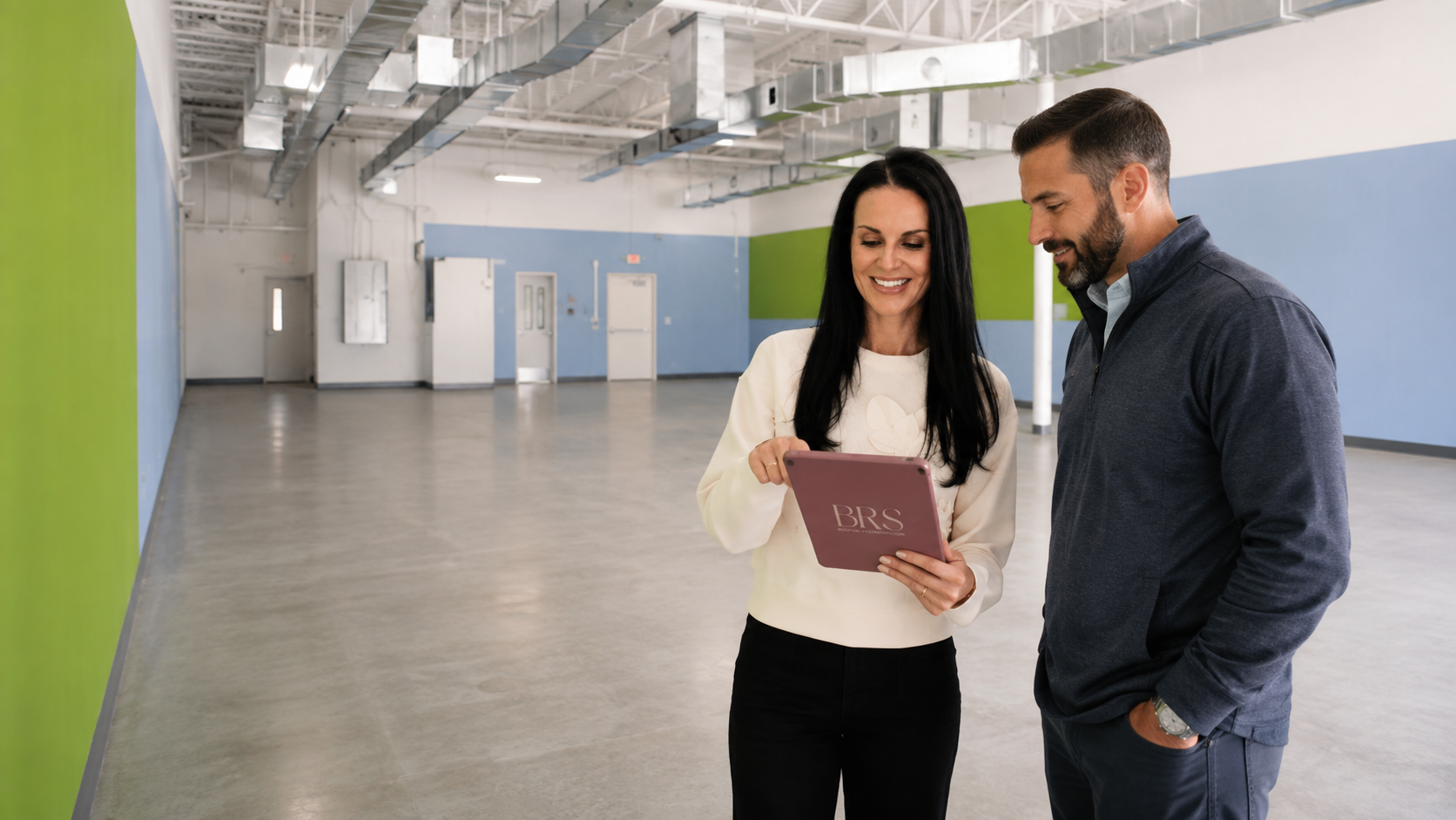 A woman and a man stand in an empty industrial or commercial space, looking at a tablet device and smiling.
