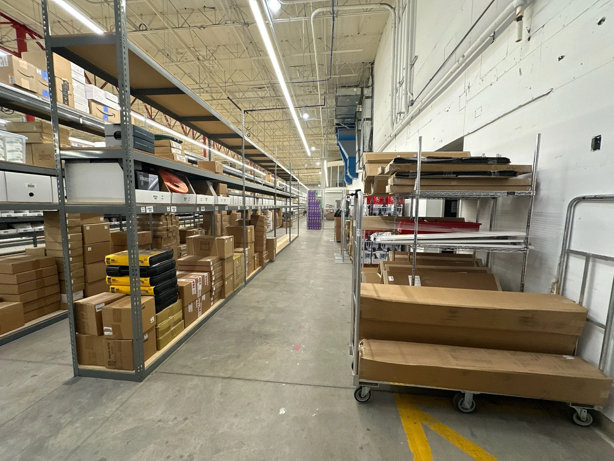 Empty retail store shelves with few items and a rolling cart stacked with empty flat boxes, located in a warehouse or large retail space.