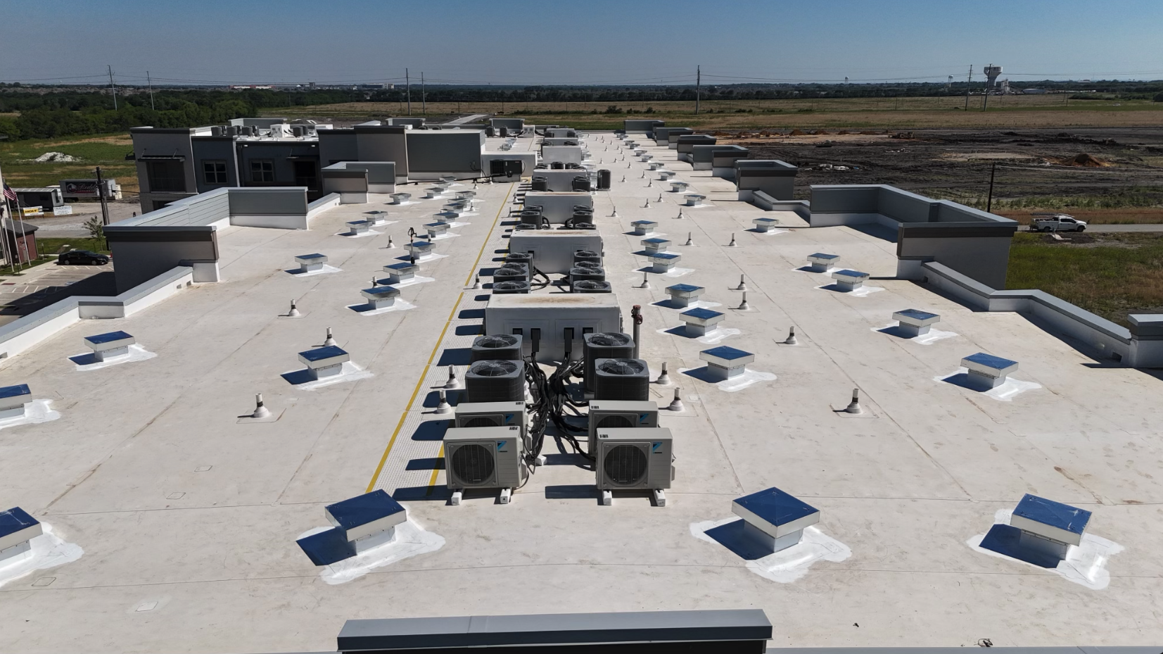 A flat commercial building roof with multiple HVAC units, vents, and skylights, overlooking a rural landscape with open fields and distant structures.