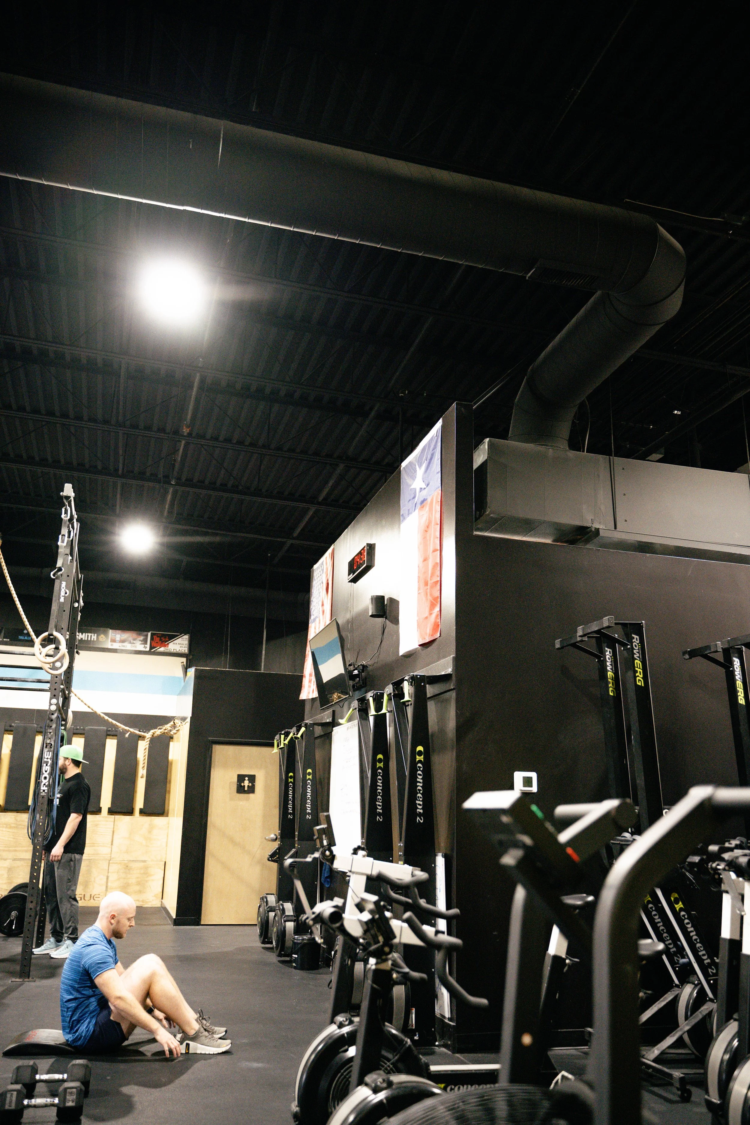 Inside a gym with various workout equipment, two men are visible. One is sitting on the floor stretching, while another stands near some equipment, both amidst a dark ceiling with bright lights and a visible ventilation duct.