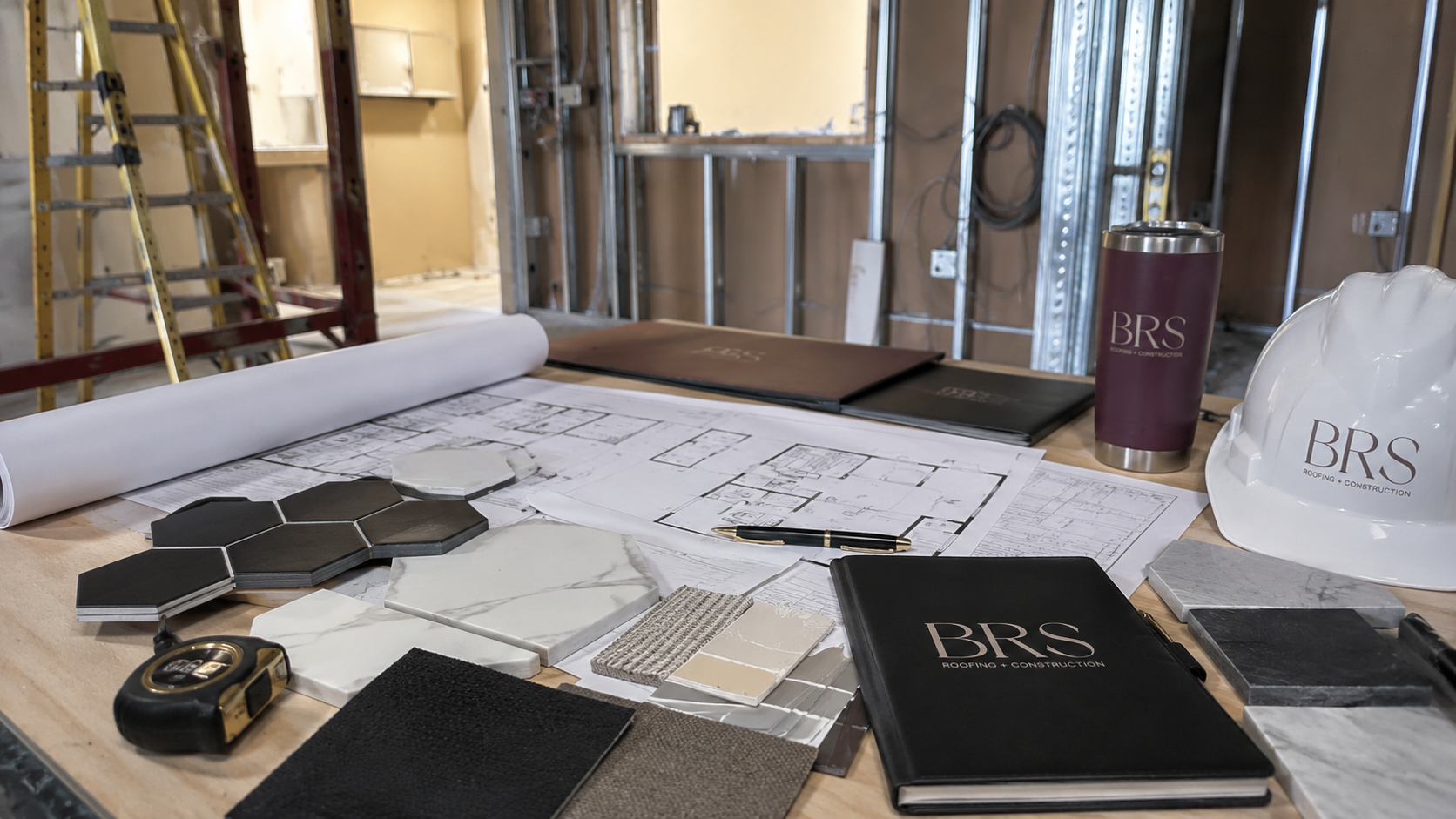 Construction planning documents and materials on a worktable, with blueprints, color samples, tiles, a measuring tape, and a safety helmet labeled 'BRS Roofing & Construction' in a building under construction.