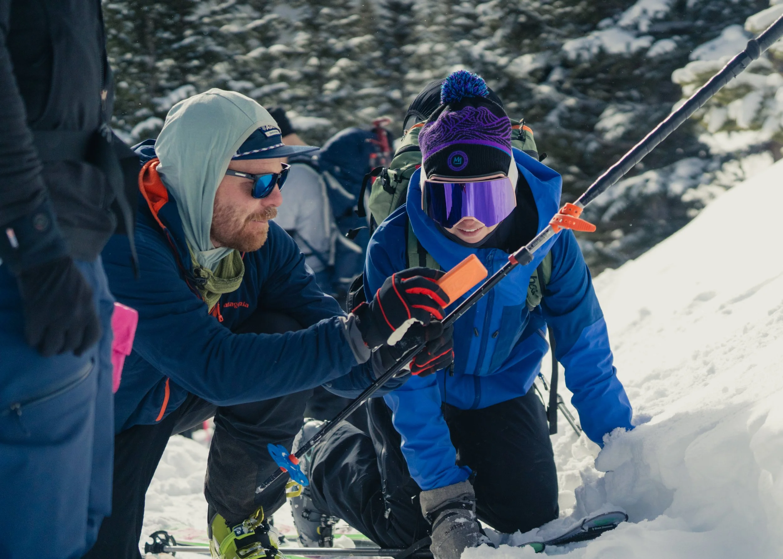 Two people dressed in winter clothing, including a hoodie and a ski jacket, are kneeling in the snow during a bright, snowy day. They are examining or working with snow gear, with one of them holding a smartphone. In the background, snow-covered tree