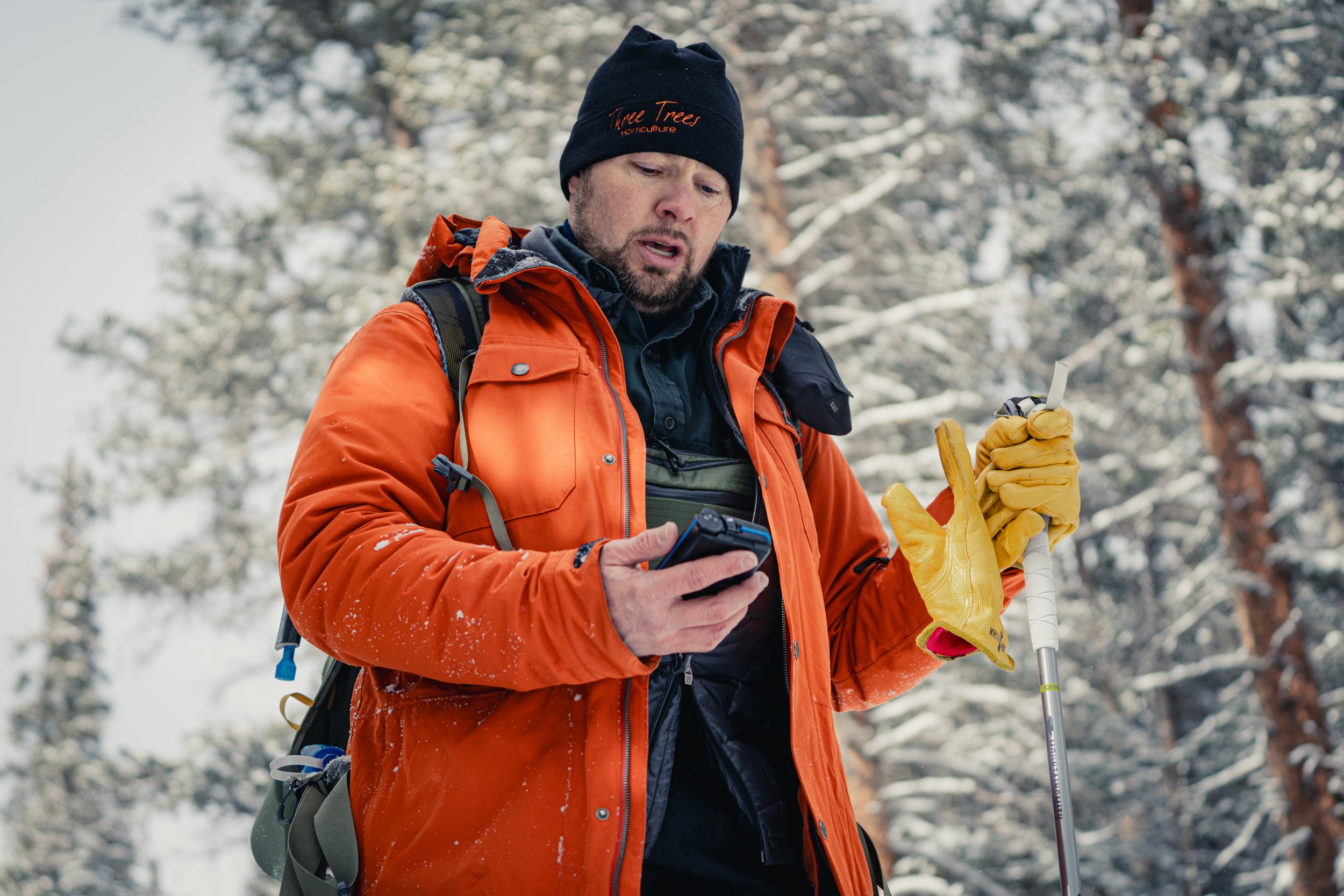A man dressed in winter outdoor gear stands in a snowy forest, holding a map or device in his right hand and a ski pole in his left, with a backpack on his back, wearing a black beanie, and an orange jacket.