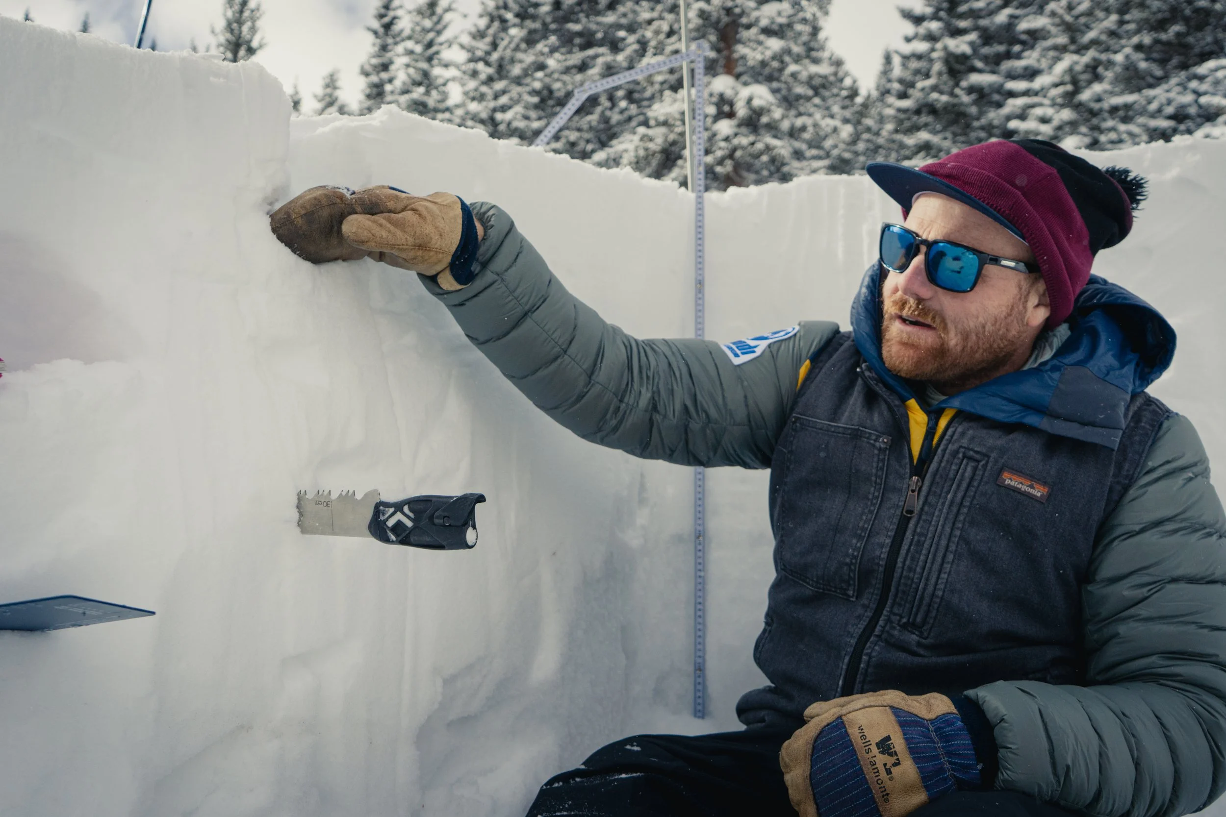 A person wearing sunglasses, a maroon beanie, gloves, and a winter jacket measures a snow depth with a ruler during a winter outdoor setting with snowy trees in the background.
