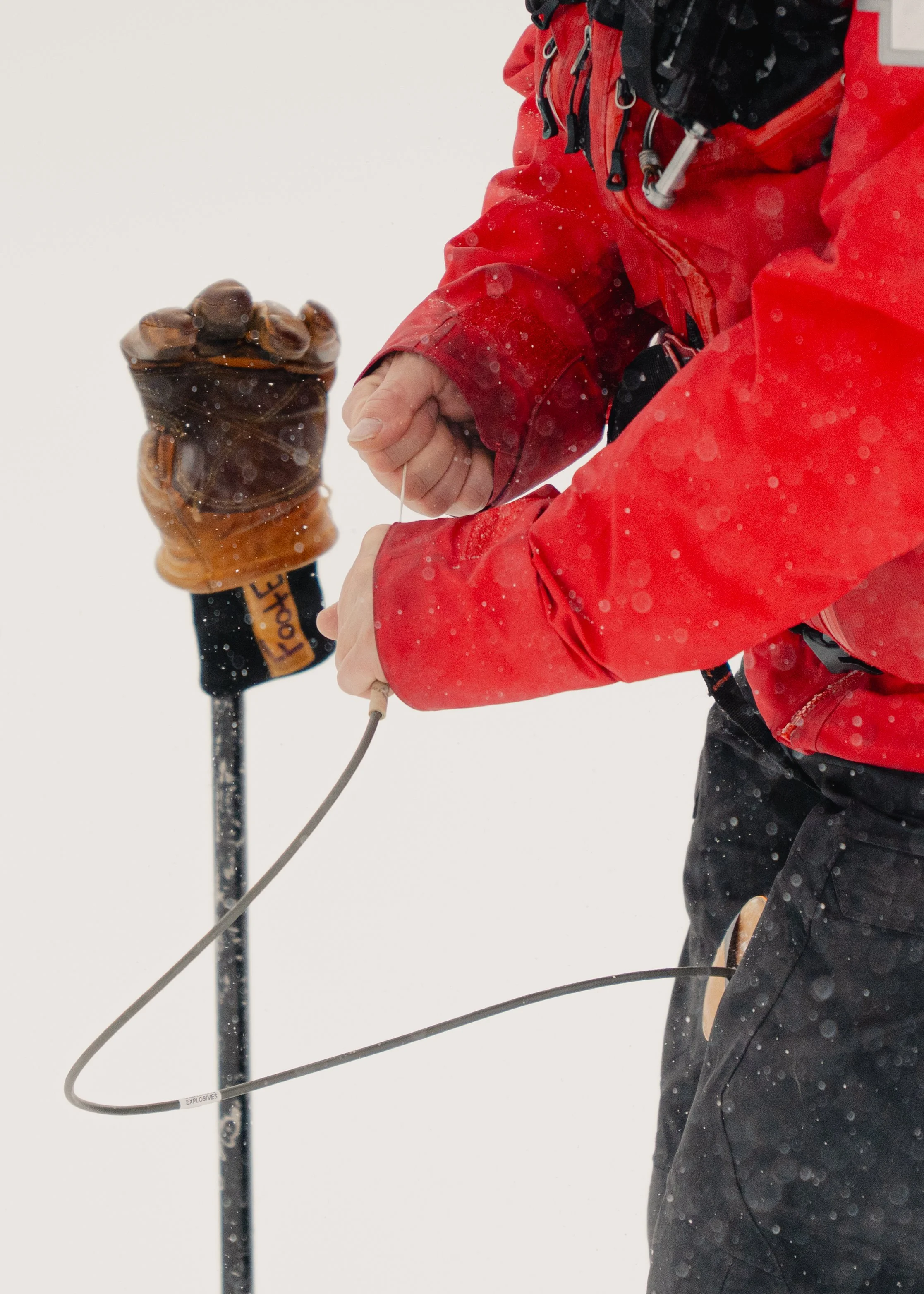 Close-up of a person in a red winter jacket, repairing or adjusting a device with a wire, with snow falling around and a snow-covered background.