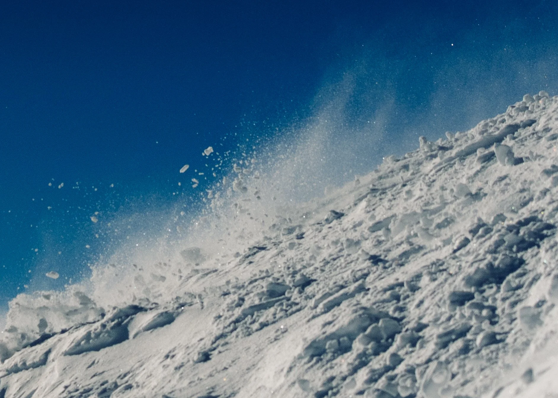 Snow-covered mountain slope with snow being blown up by the wind against a clear blue sky.