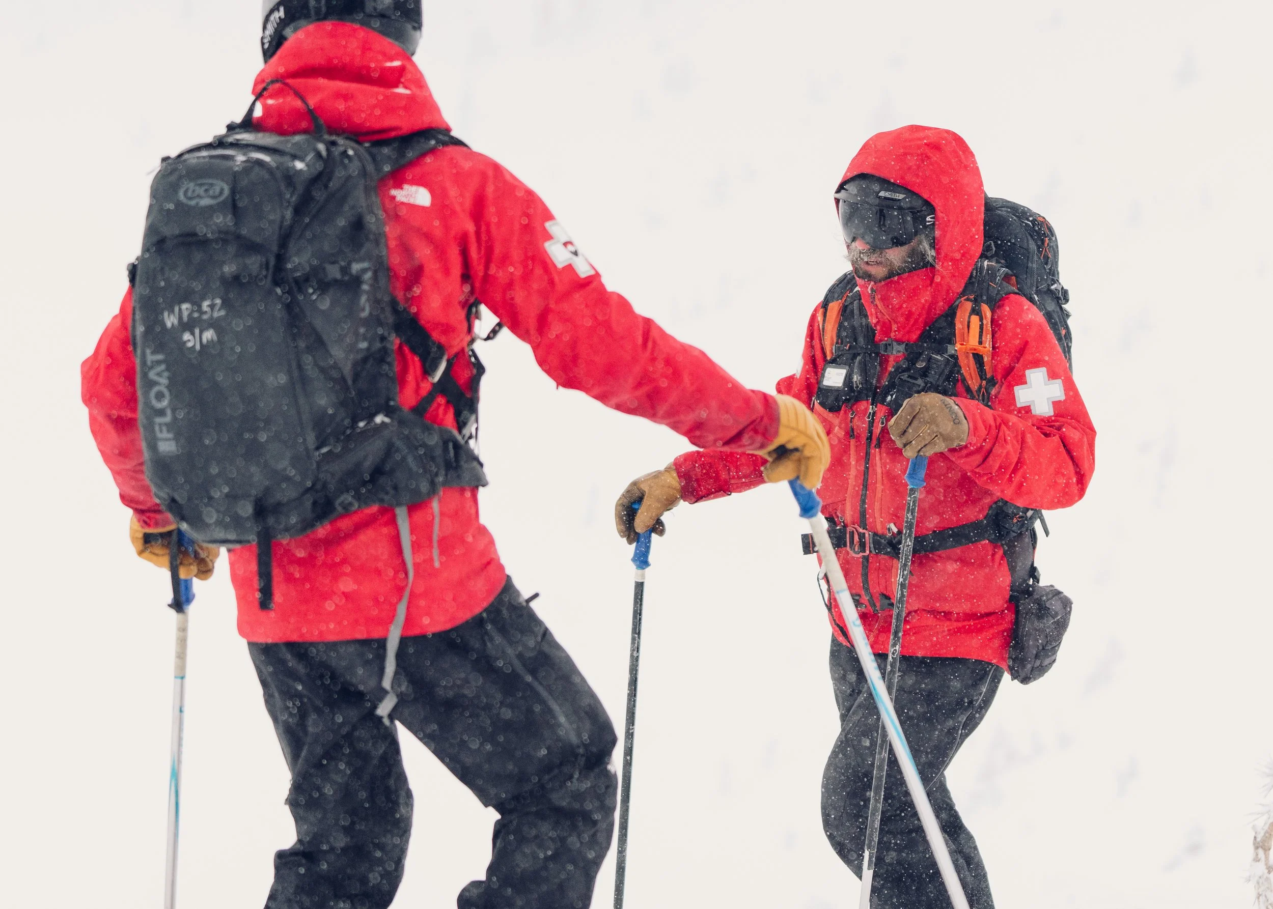 Two people dressed in red winter gear and backpacks standing in snowy weather, facing each other, with one of them holding ski poles.