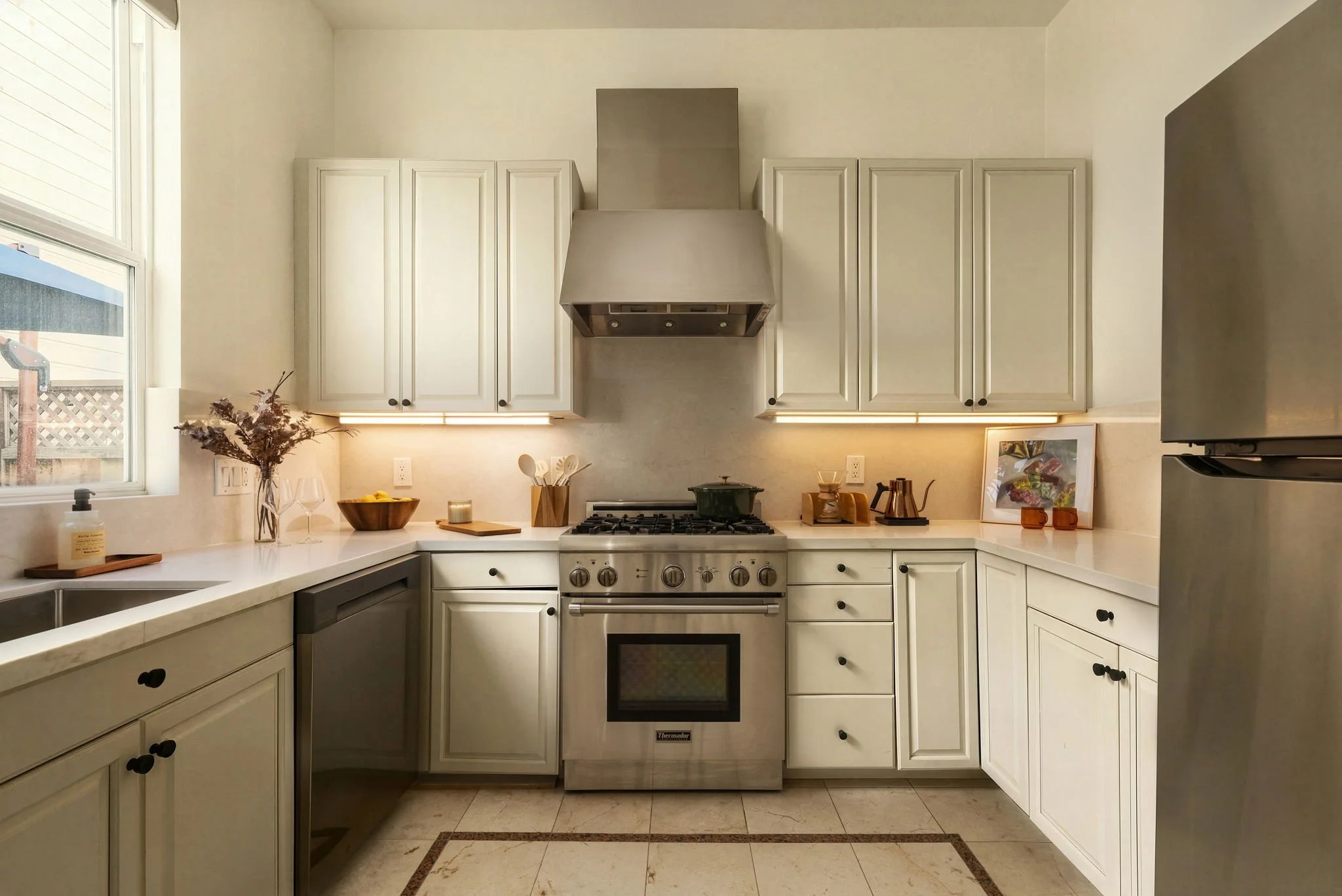 A modern kitchen with white cabinets, a stainless steel range hood above a gas stove, a black oven, and various kitchen items on the countertop, including a bowl of lemons, a vase with dried flowers, and framed artwork.
