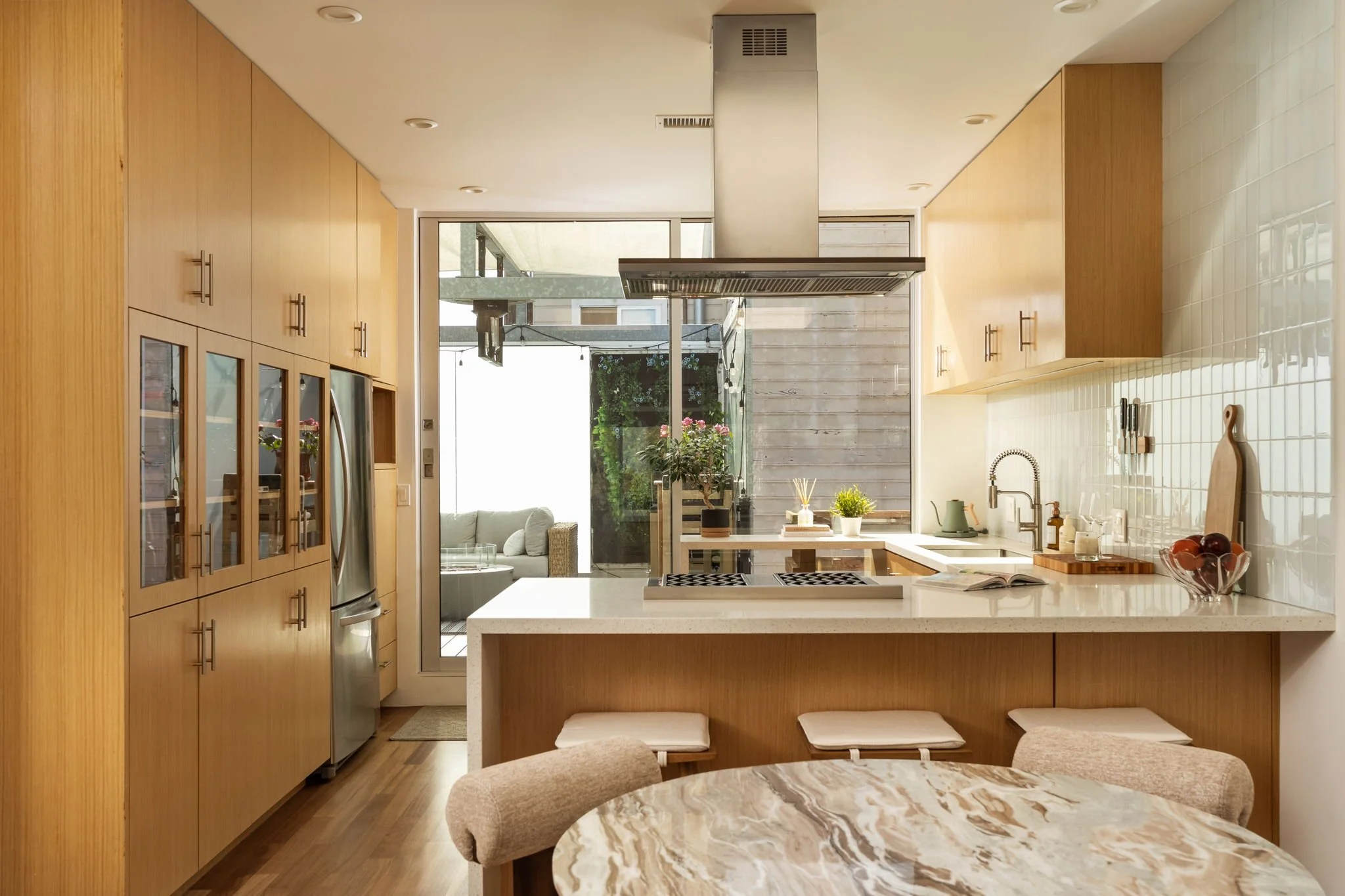 Modern kitchen with wooden cabinets and white countertops, view of a backyard with garden and outdoor seating visible through a glass door.