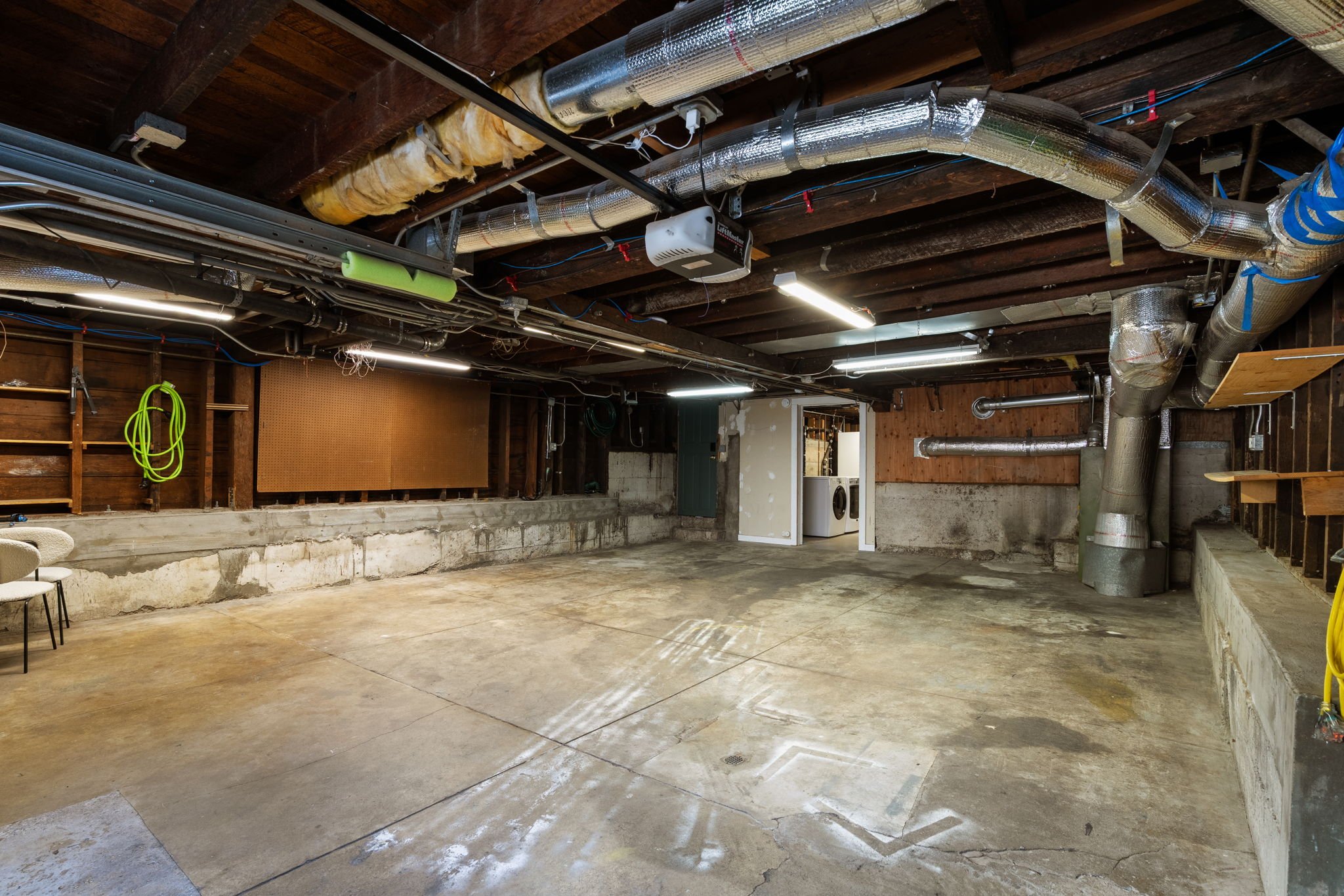 A basement with exposed wooden ceiling and ducts, a washer and dryer, unfinished concrete walls and floor, and partially installed wall panels.