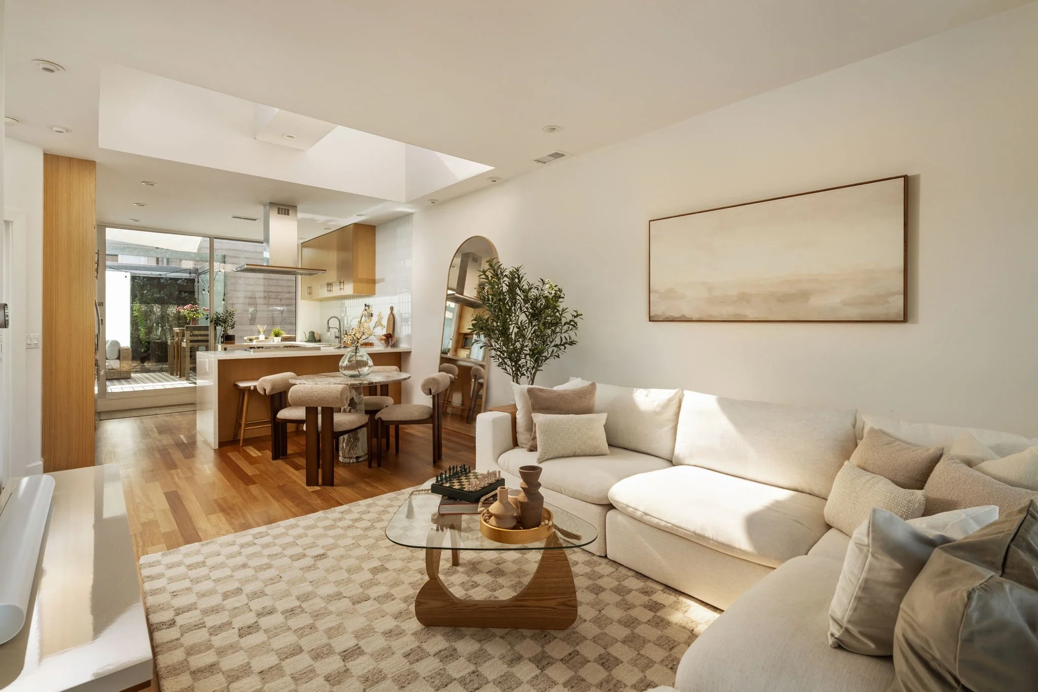 Bright, spacious living room with white walls, wooden flooring, a large white sofa with beige and white pillows, a glass coffee table with decorative vases, and a large artwork on the wall. Adjacent is a dining area with a wooden table, chairs, and a