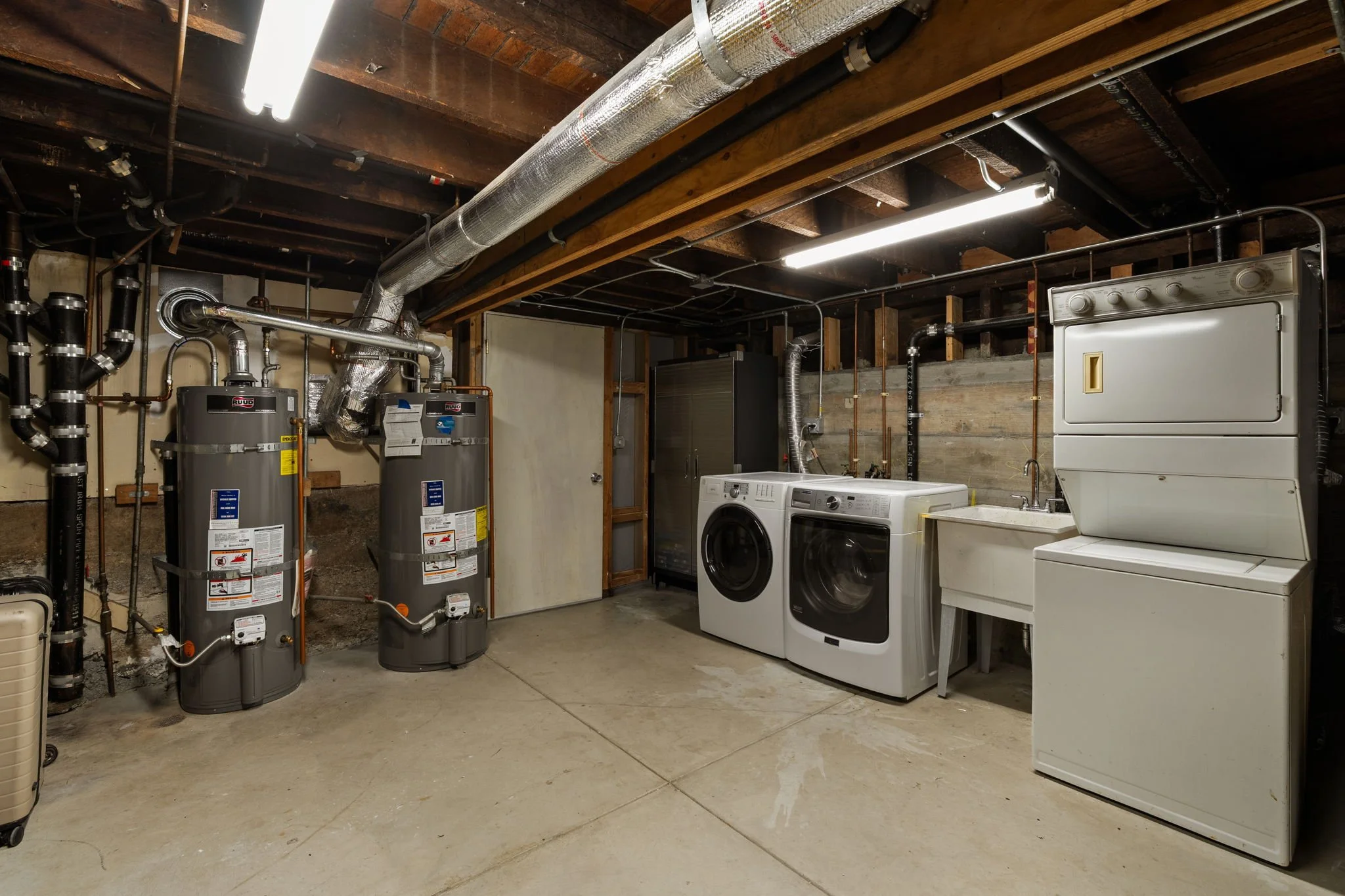 A laundry room with a washing machine, a dryer, water heaters, and exposed ceiling pipes and wooden beams in an unfinished basement.