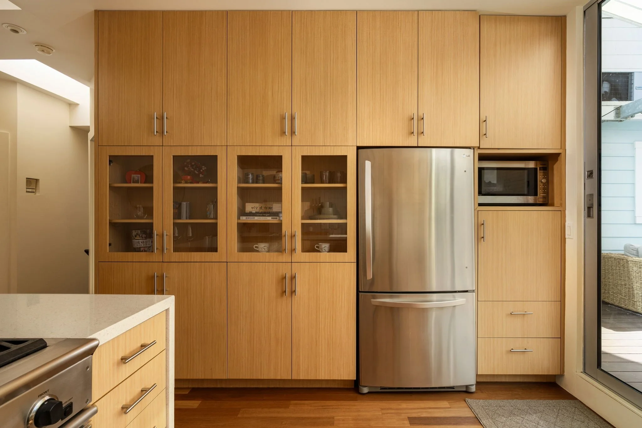 Kitchen with wood cabinets, a stainless steel refrigerator, and a microwave oven built into the cabinetry. Part of a countertop is visible in the foreground. A glass door on the right opens to an outdoor space.