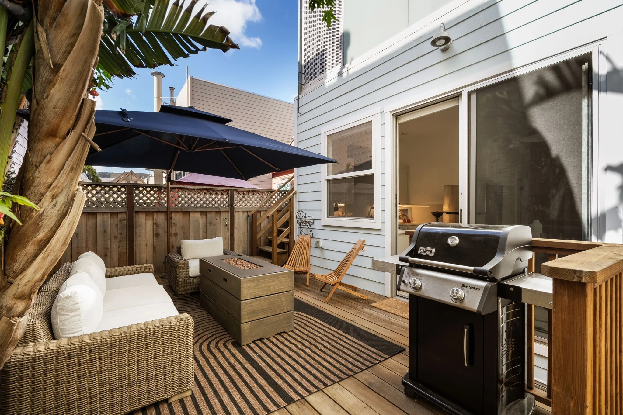 A backyard patio area with outdoor furniture, large black umbrella, barbecue grill, and wooden deck, surrounded by a white house and a wooden fence, with a banana tree in the foreground.