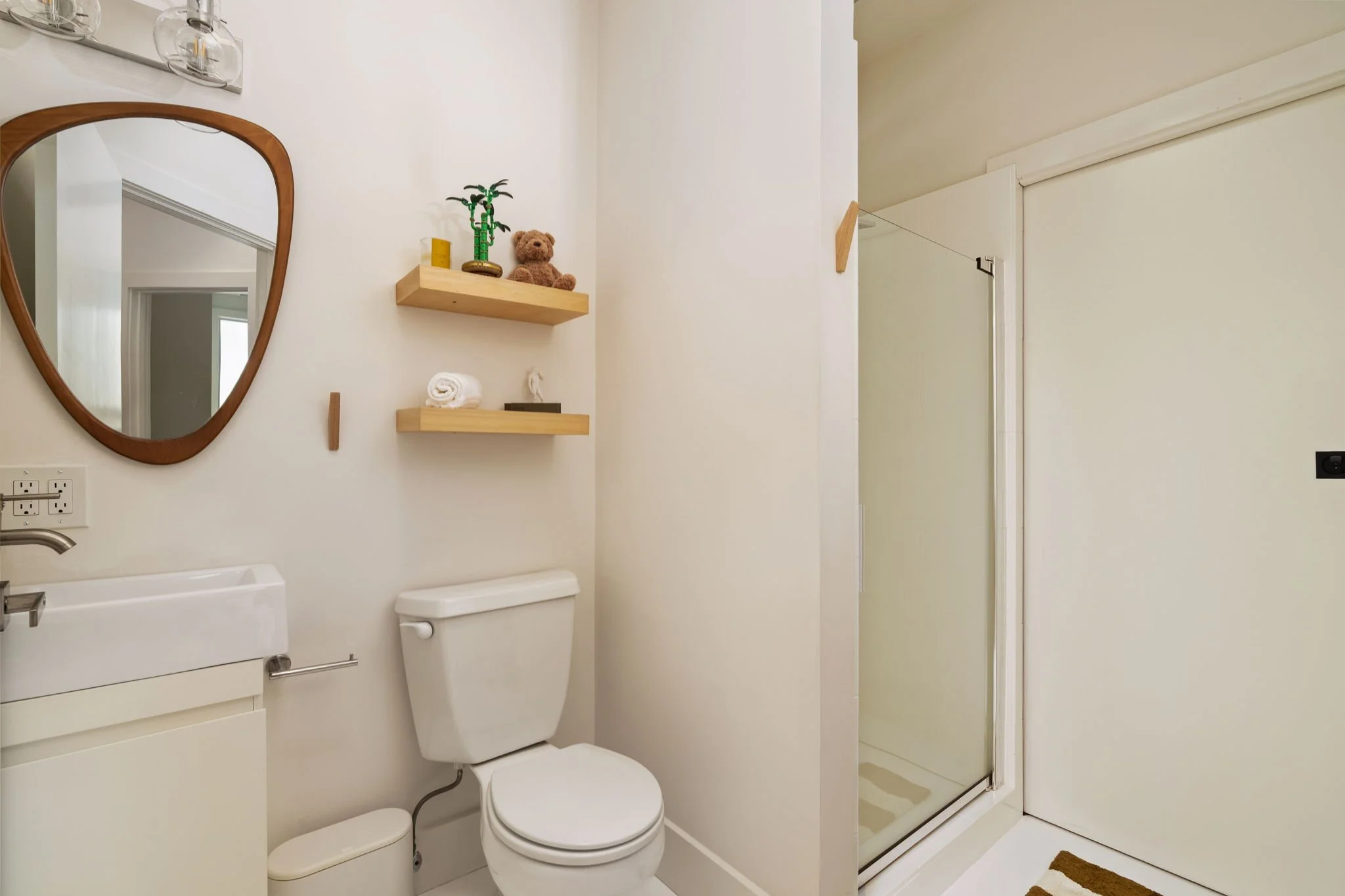 A modern bathroom with a white toilet, a cabinet below the sink, a mirror with a wooden frame, two wooden shelves with decorative items, a shower with a glass door, a brown rug, and a white wall and door.