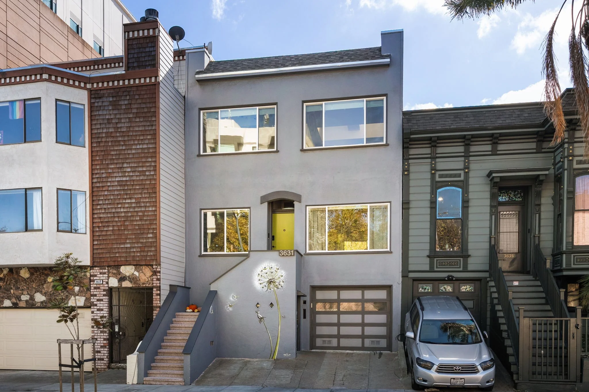 A row of houses with diverse architectural styles in an urban neighborhood. The central house is a modern, gray, rectangular structure with large windows, gray front stairs, and a yellow front door. To the right, an older, Victorian-style house featu