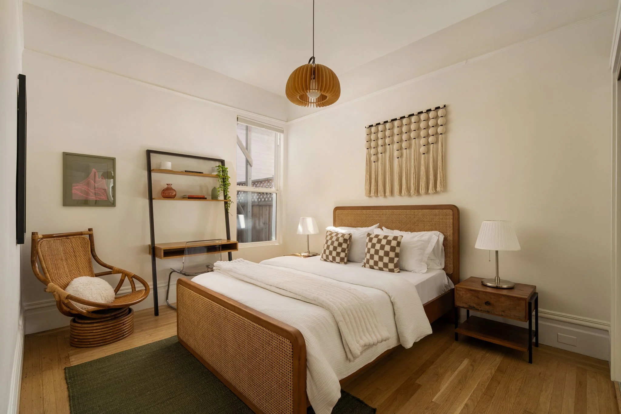 A bedroom with a bed, patterned pillows, two table lamps, wicker chair, wall decorations including a wooden wall hanging and a framed art piece, and a window with a view of a wooden fence outside.