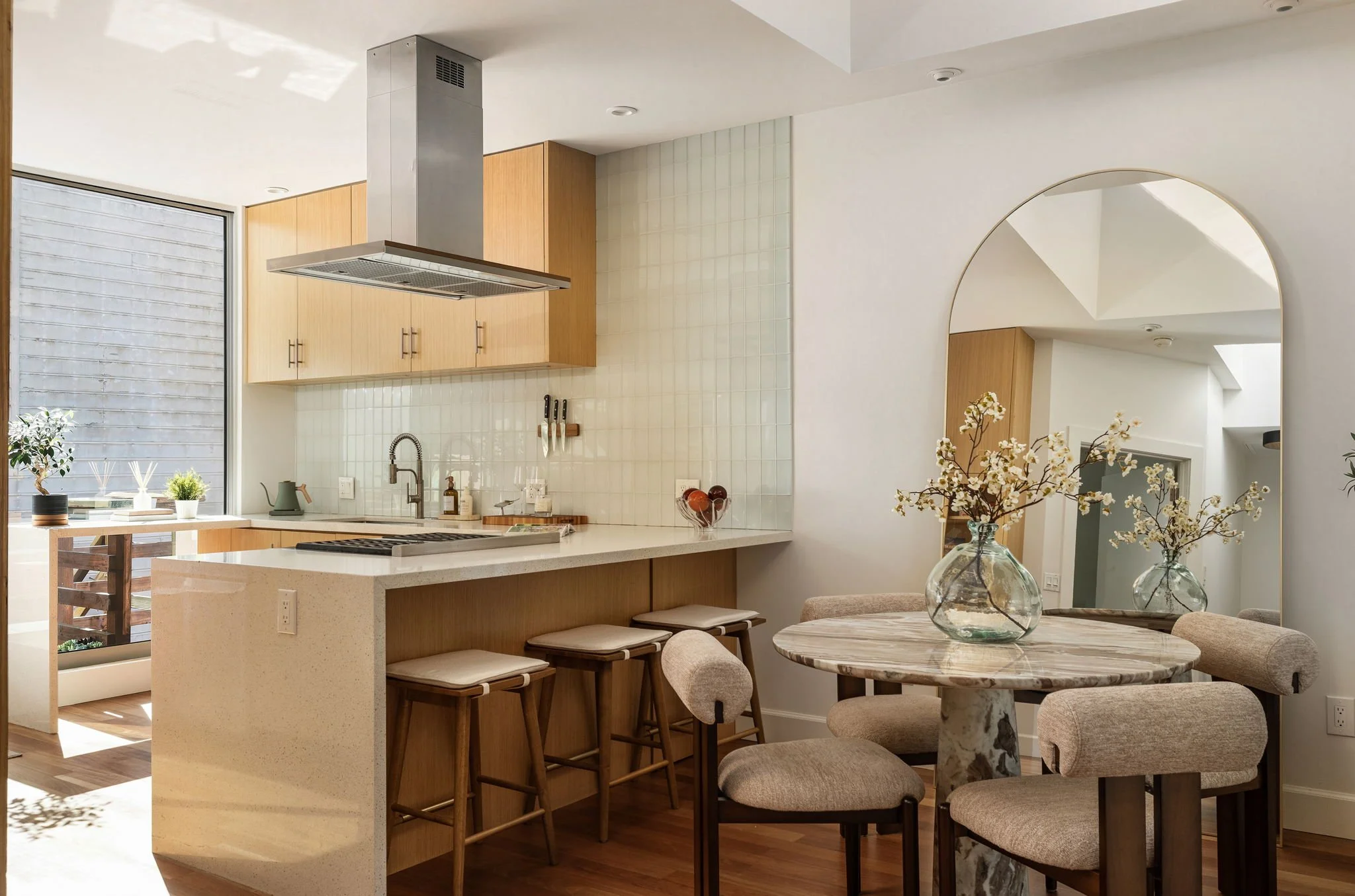 Modern kitchen with wooden cabinets, white countertops, a hanging range hood, and a dining area with beige upholstered chairs and marbled vases with flowers.