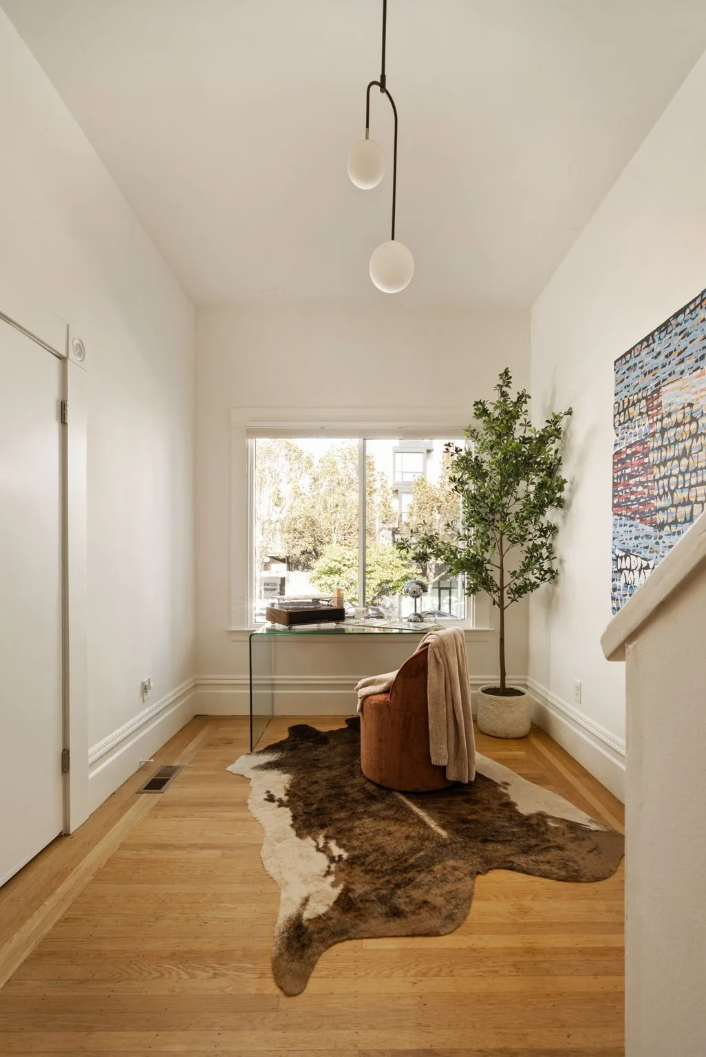 A home office corner with a window, a brown upholstered chair with a blanket, a glass desk with some items, a tall potted plant, an abstract wall art piece, and a cowhide rug on hardwood floors.