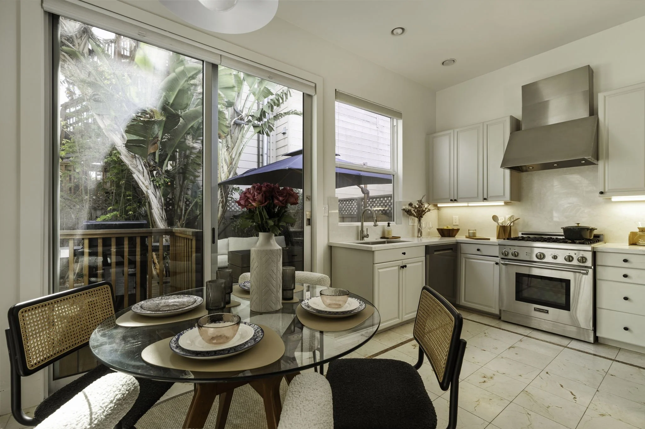 Interior view of a kitchen and dining area with a glass-top table, chairs, white cabinets, a stove, and sliding glass doors leading to a backyard patio with an umbrella and plants.