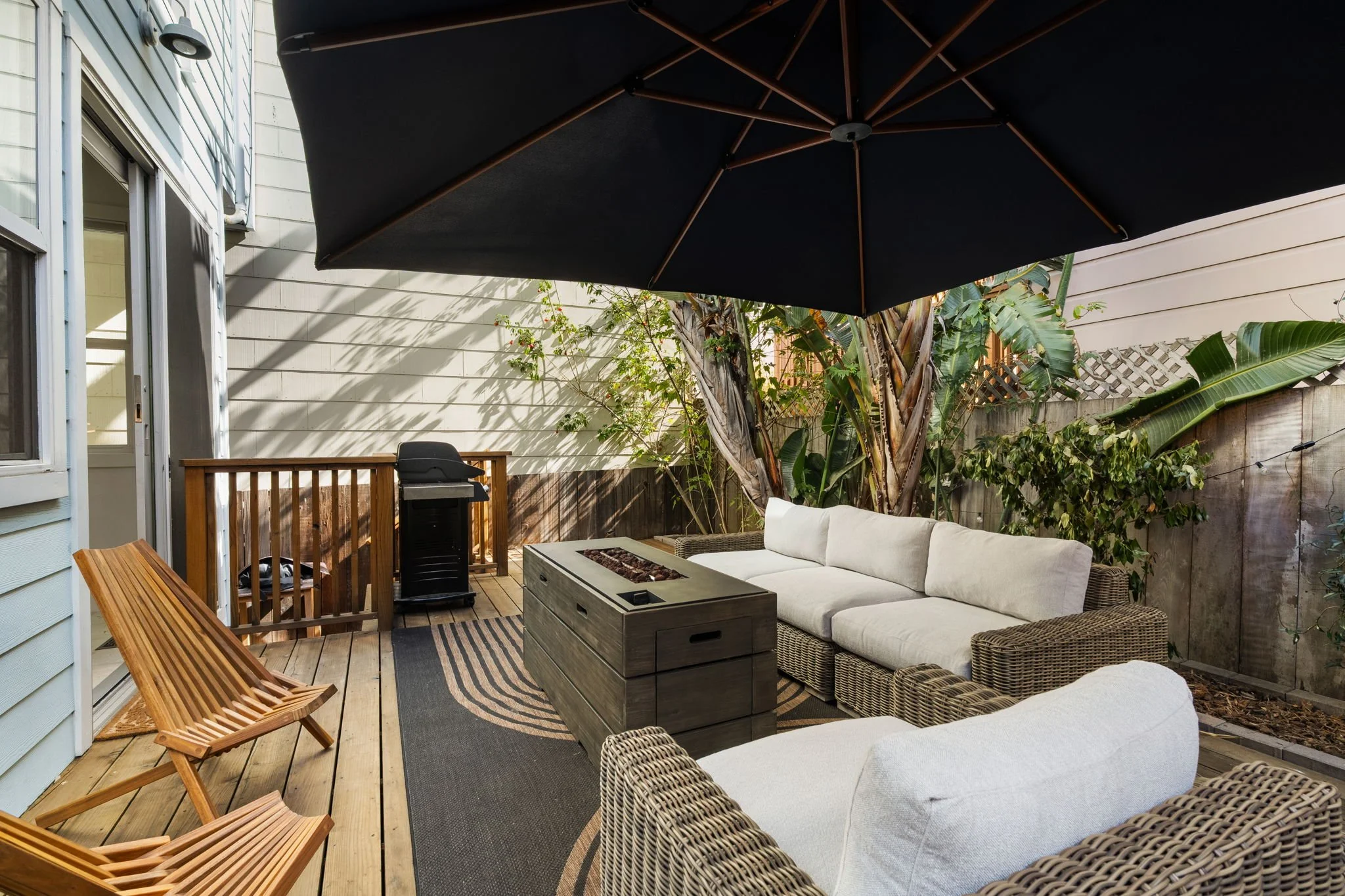 An outdoor patio with wicker and wooden furniture, a large black umbrella, a white sectional sofa, a fire pit table, a small grill, and lush green plants in a backyard setting.