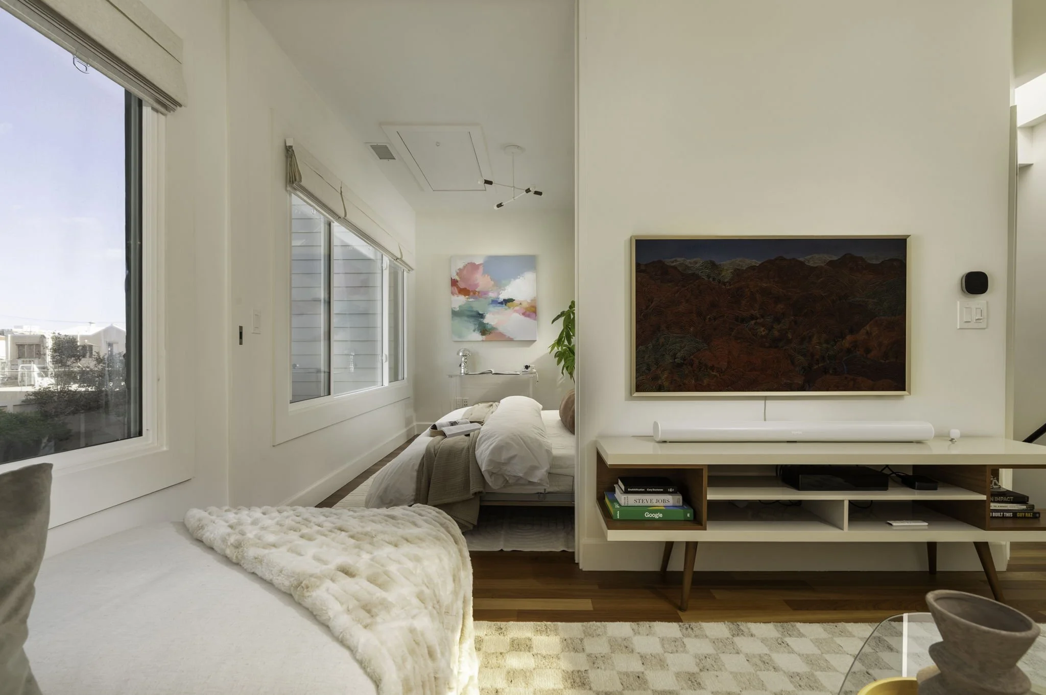 A bright living room with large windows, white walls, and wooden flooring. There are two framed artworks on the wall and a white couch. A TV sits on a white entertainment unit below the larger artwork, with several books and decorative items on the s