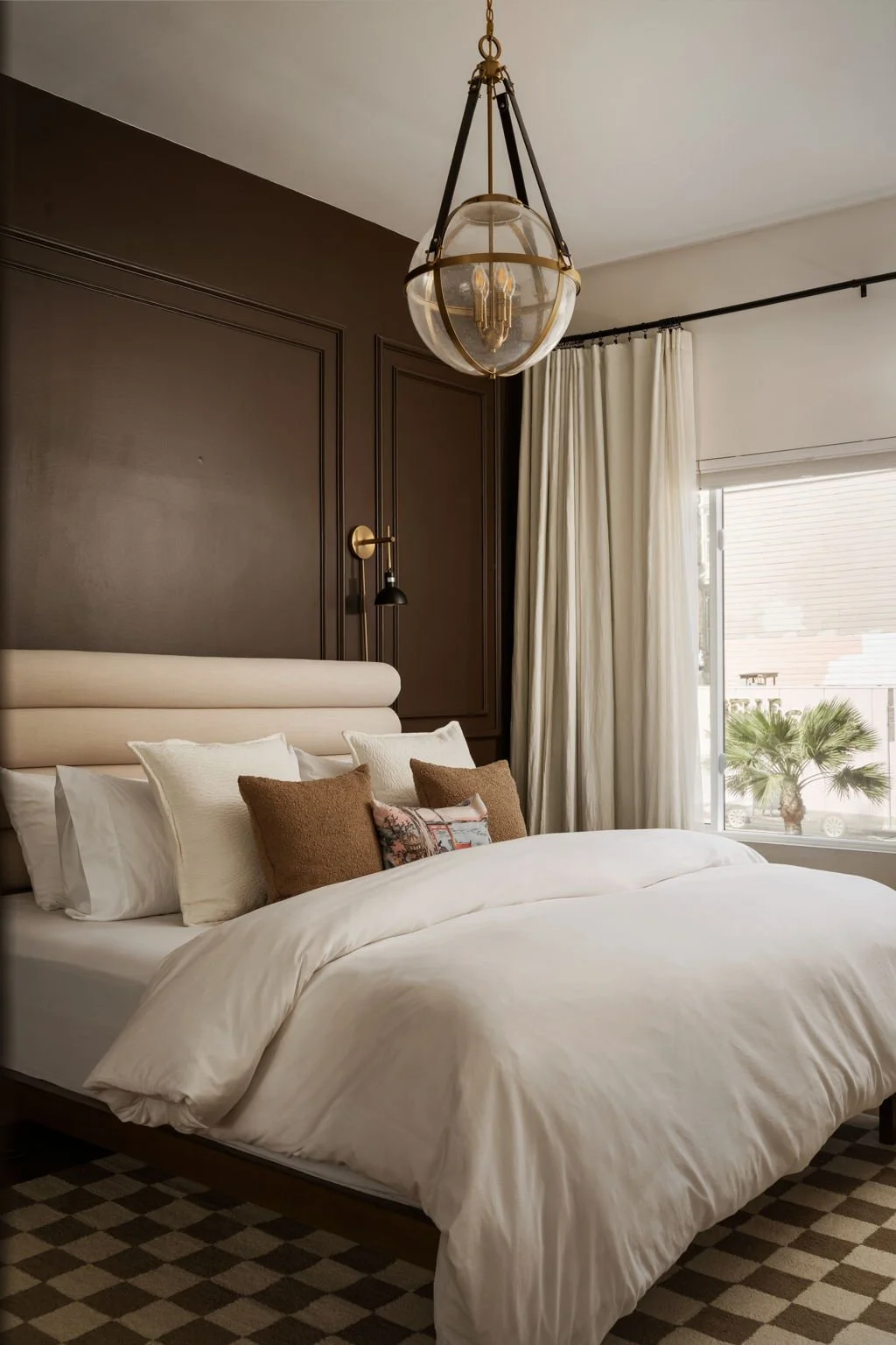 A neatly made bed with white bedding and multiple pillows, set against a dark accent wall in a well-lit bedroom with large window and white curtains.
