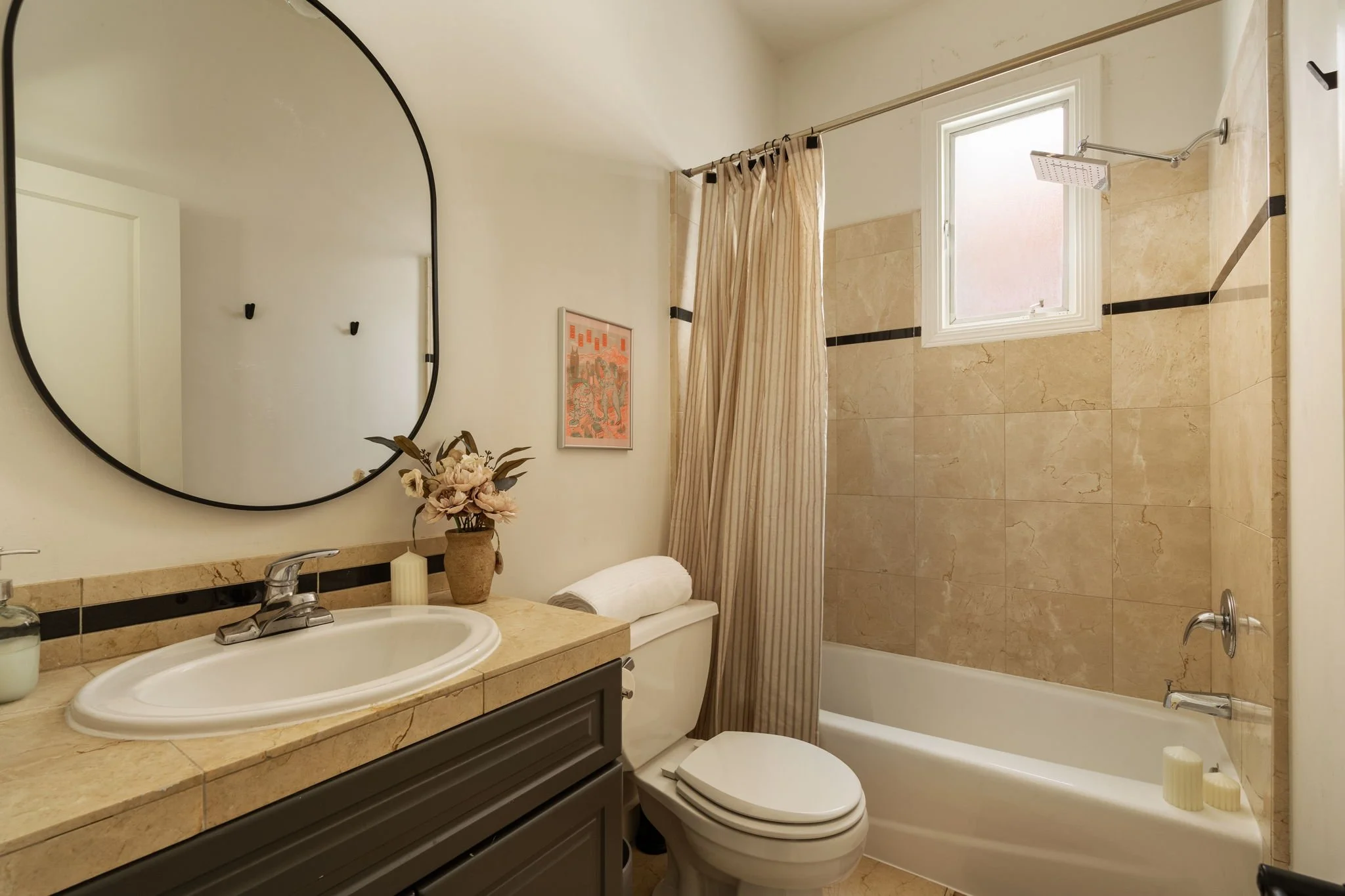 A bathroom with a beige tiled wall, a window, a shower with a curtain, a white bathtub, a vanity with a sink, a mirror, a vase with flowers, and some decorative items.