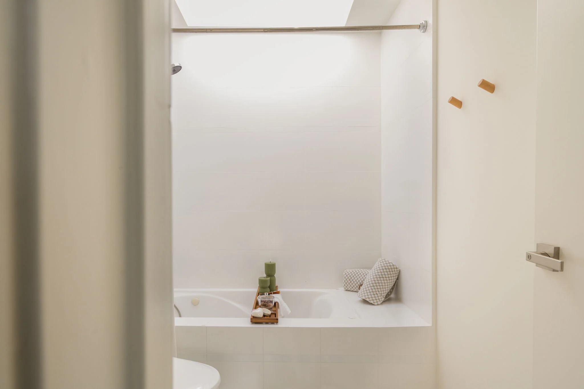 View of a modern bathroom with a bathtub, a showerhead, and decorative wooden hooks on the wall. Items like a wooden tray with bath accessories, pillows, and candles are placed around the bath area.