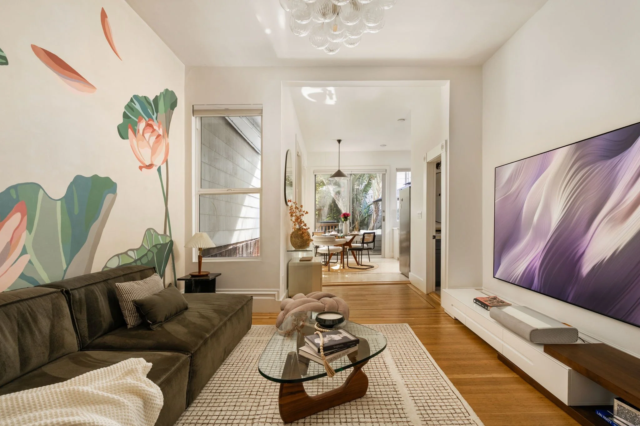 Living room with a dark gray sofa, a glass coffee table, and a large flat-screen TV on the wall. The walls are white with colorful floral wall art, and there is a round dining table with chairs visible through an open doorway. Natural light enters th