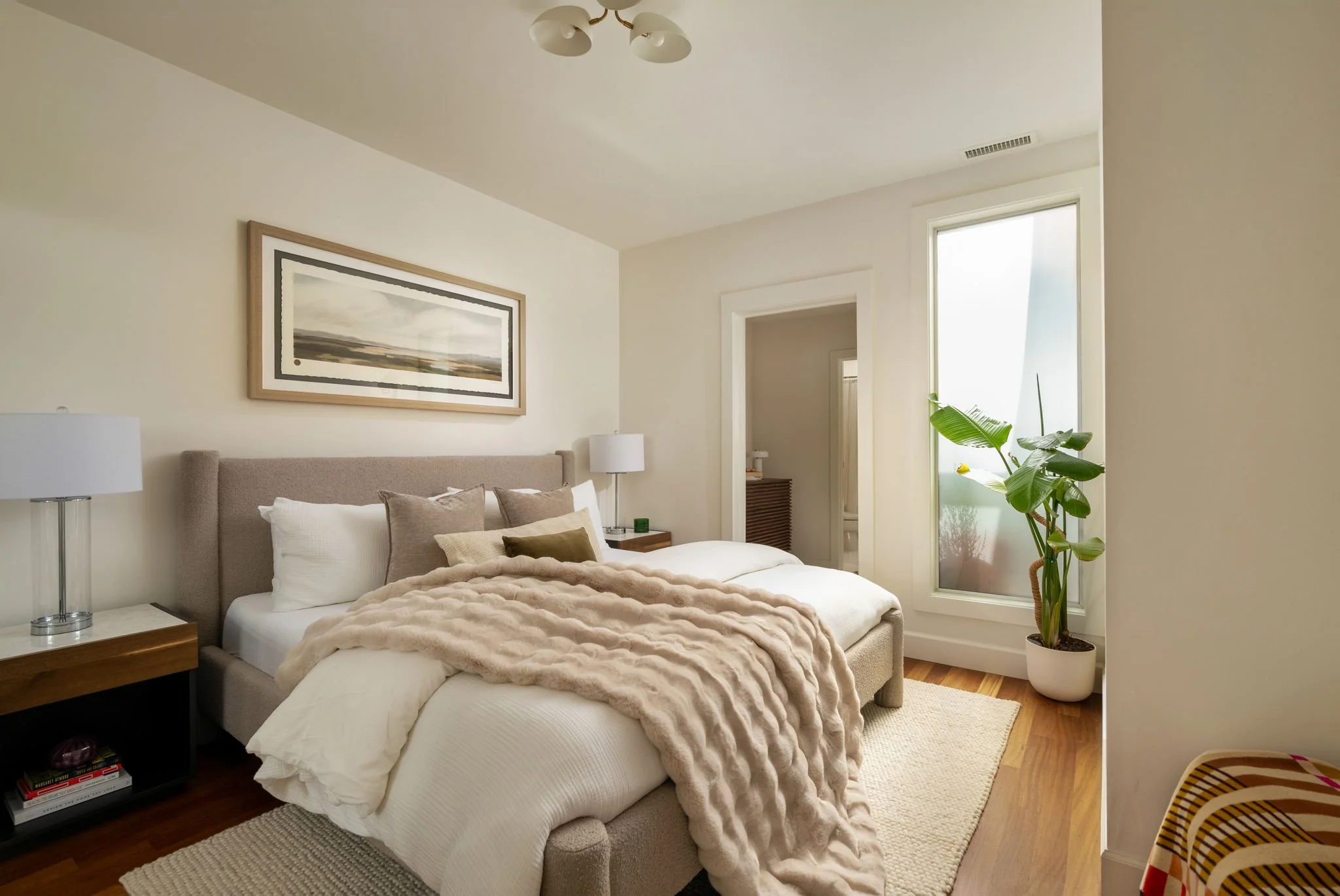 A cozy bedroom featuring a large bed with white and beige pillows and a fur blanket, two bedside tables with white lamps, a framed landscape picture on the wall, a tall window with sunlight, a potted plant, and a striped chair.