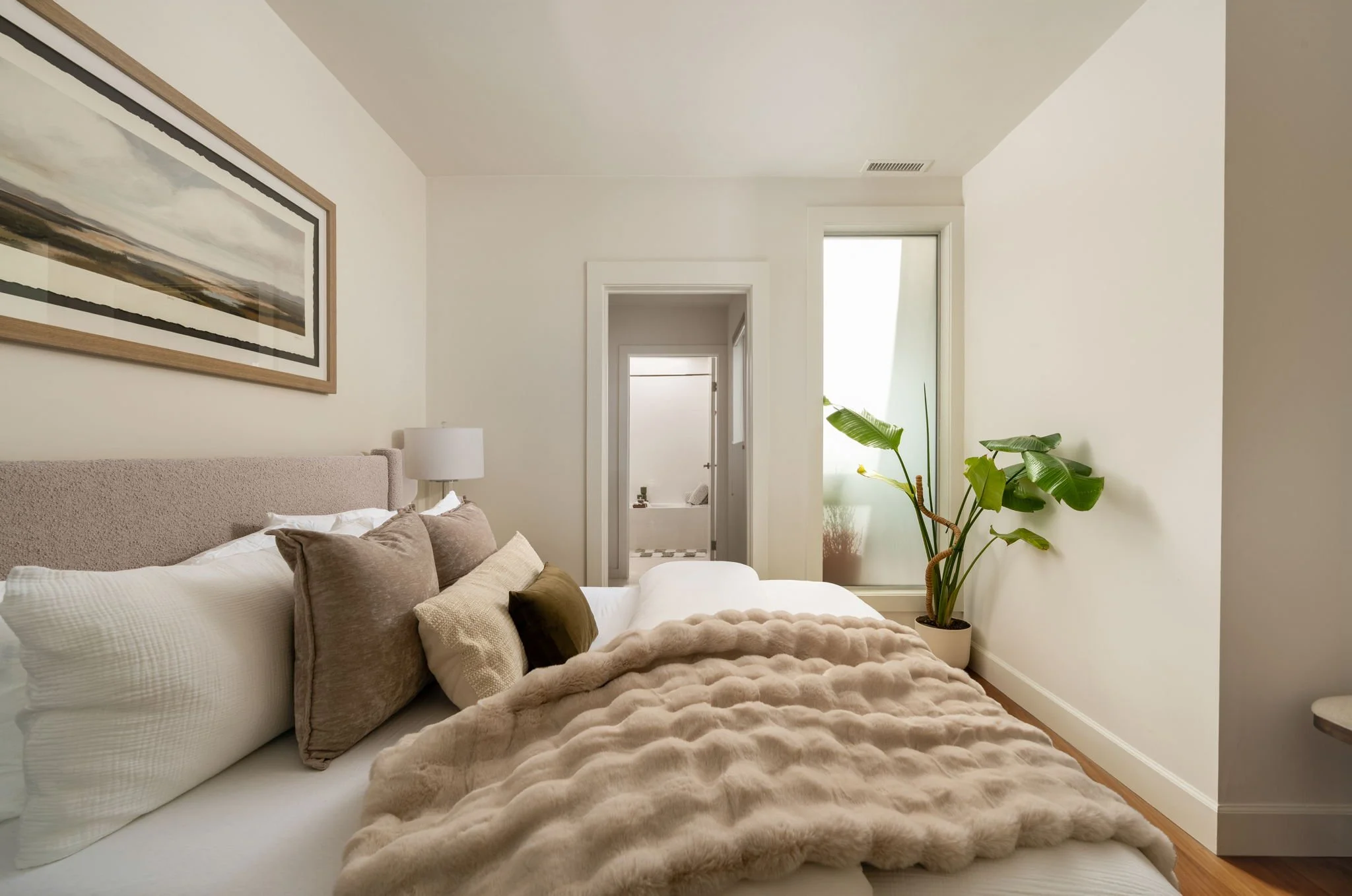 A cozy bedroom with a large bed covered in a plush beige blanket and multiple pillows, a framed landscape painting on the wall, a bedside lamp, and a tall green plant near a window. The walls are painted white, and there are doorways leading to other