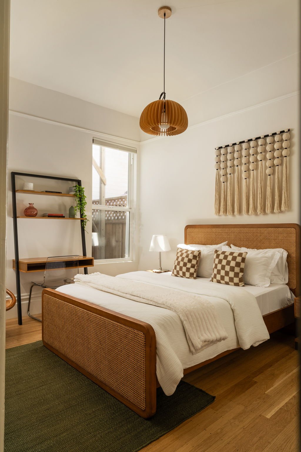 A cozy bedroom with a wooden bed frame, white bedding, checkered pillows, a bedside lamp, a wall hanging with tassels, a window with natural light, a small desk with a chair, and a black shelf with decorative items and a plant.