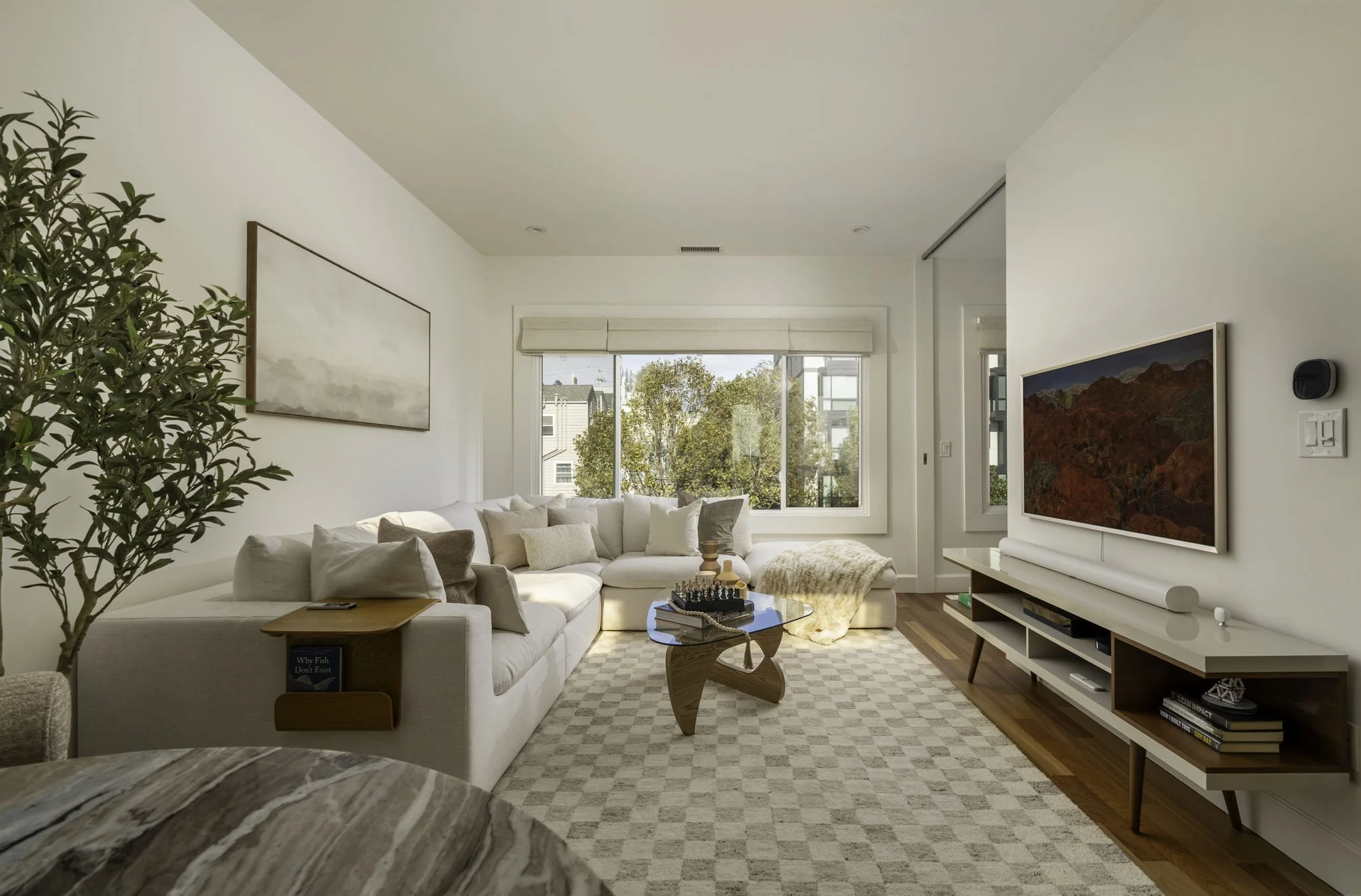 Bright living room with white walls, large window, cream sectional sofa with pillows, wooden coffee table with chess, wall art, greenery, and TV.