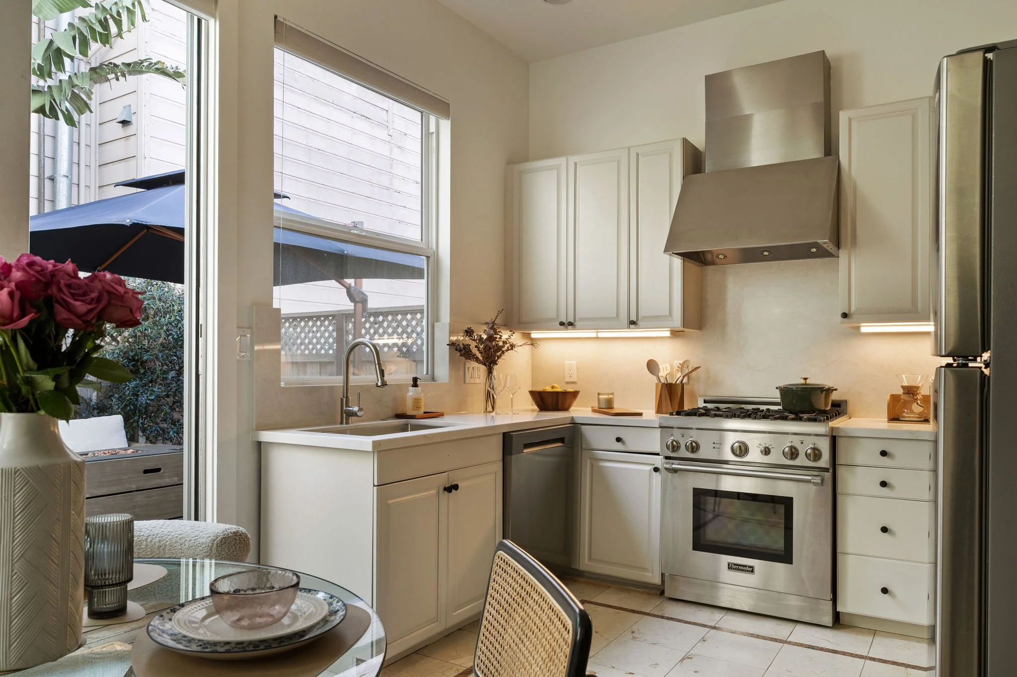 A modern kitchen with white cabinetry, stainless steel appliances, and large windows letting in natural light. There is a table with a vase of pink roses and glassware.