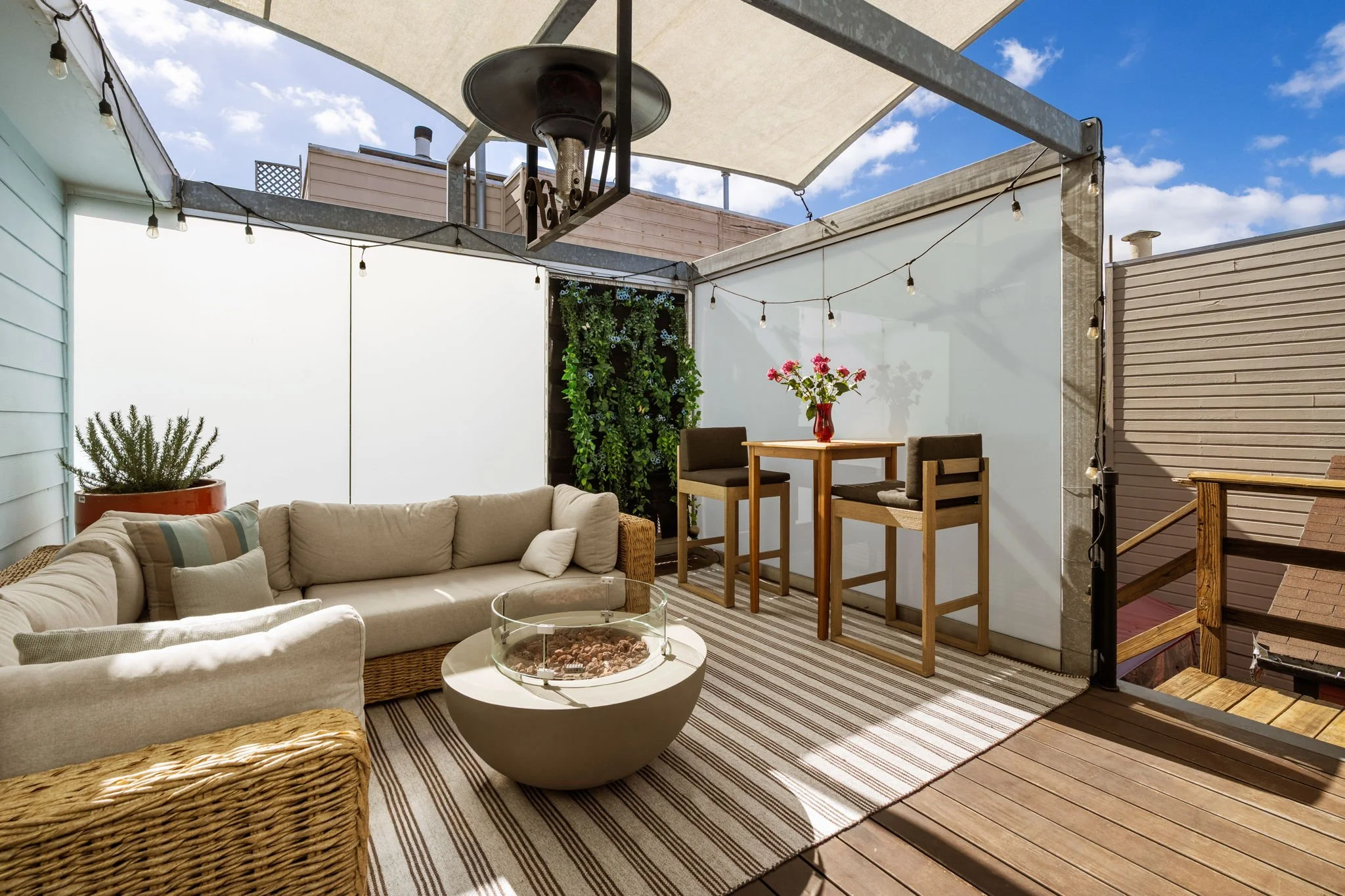 Outdoor patio with a beige sectional sofa, wicker chair, wooden high-top table with two chairs, decorative plant wall, potted plant, string lights, striped outdoor rug, wood deck, and a white sunshade on a sunny day.