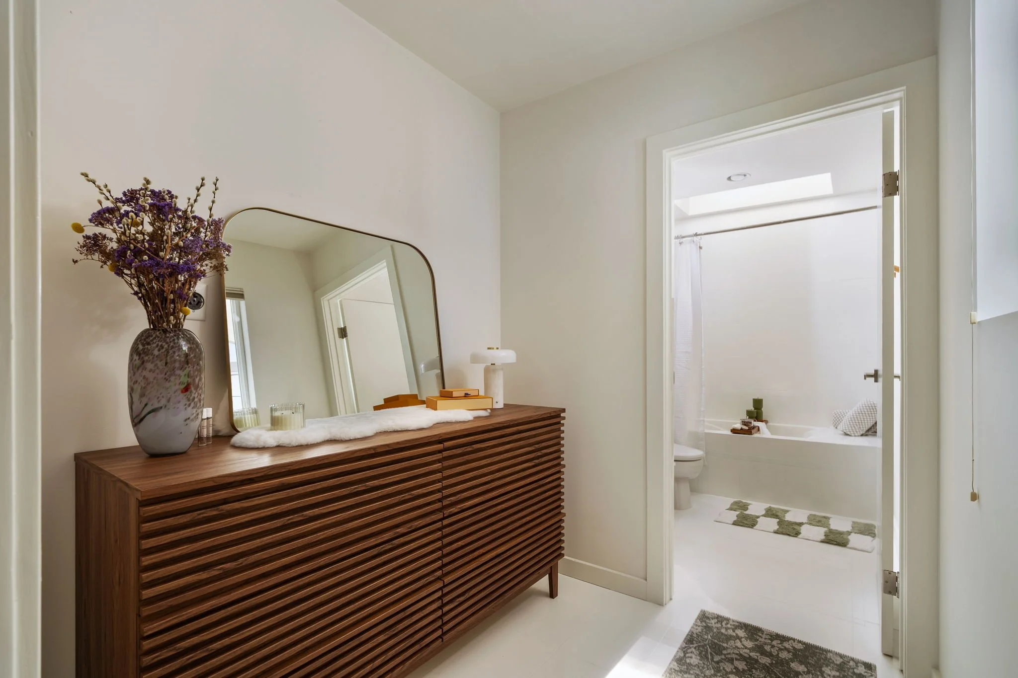 A bathroom with a wooden dresser, a vase with purple flowers, a mirror, and a small table with a lamp and books. The doorway reveals a tub and a shower curtain.