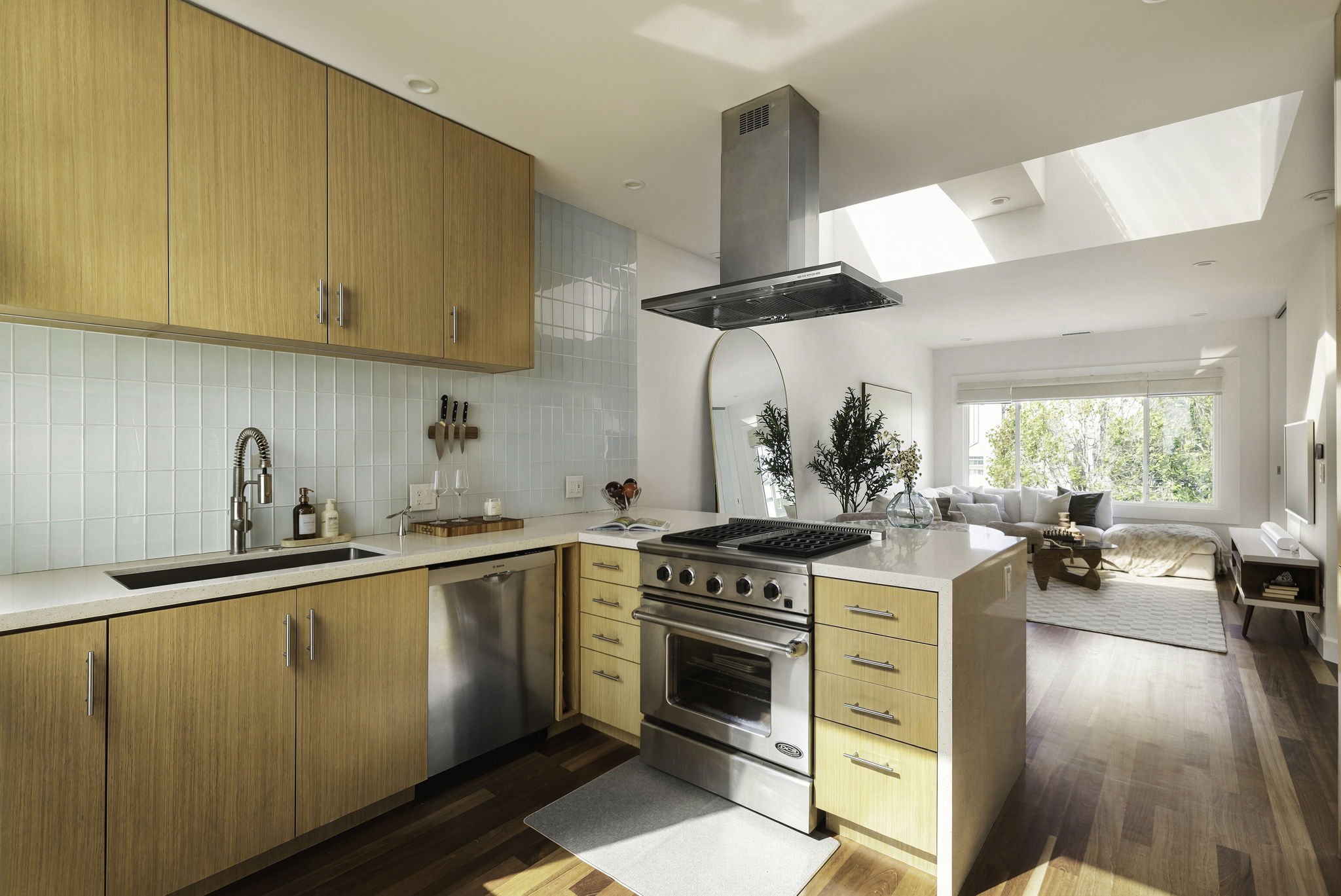 A modern kitchen with light wood cabinets, a white countertop, a stainless steel stove, and a light green tiled backsplash. The kitchen opens to a living room with large windows, a white sofa, and indoor plants.